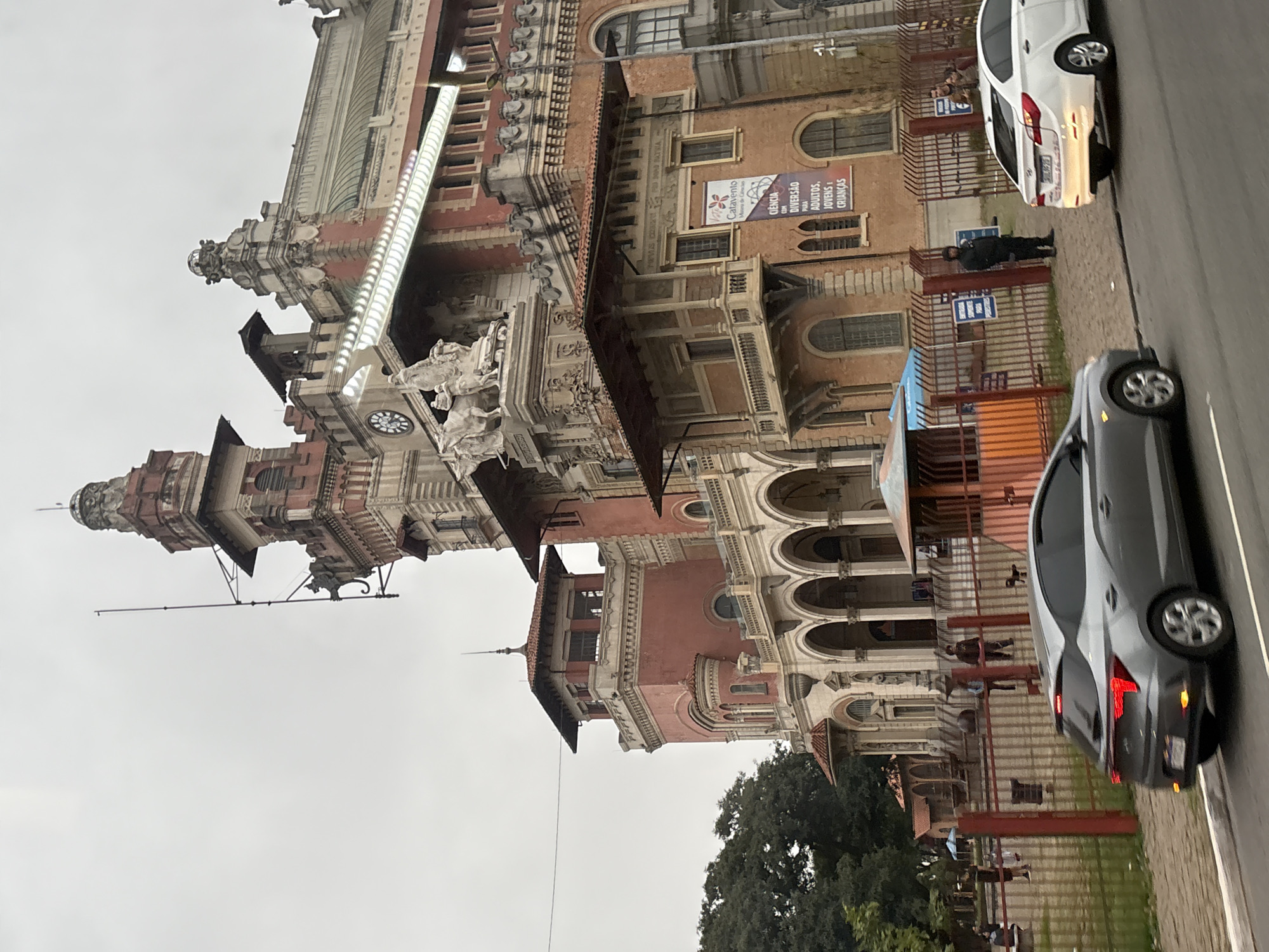 The image shows the exterior of a large, ornate building with a red and pink facade, featuring multiple towers, arches, and decorative elements. This building is the Museu de Zoologia da USP (Museum of Zoology of the University of São Paulo), located in the Ipiranga district of São Paulo, Brazil.
In the foreground, a gray hatchback car is driving on a road, and a white sedan is parked on the side of the road. A metal fence separates the sidewalk from the museum grounds. Behind the fence, people are visible walking around the museum's exterior and in a park-like area.
The sky is overcast, and the overall lighting suggests it is daytime. The architecture of the museum is a prominent feature, with its classical and baroque influences. There are banners and signs displayed on the museum's walls. A security guard is standing near the fence. The environment appears to be a public space within the city.