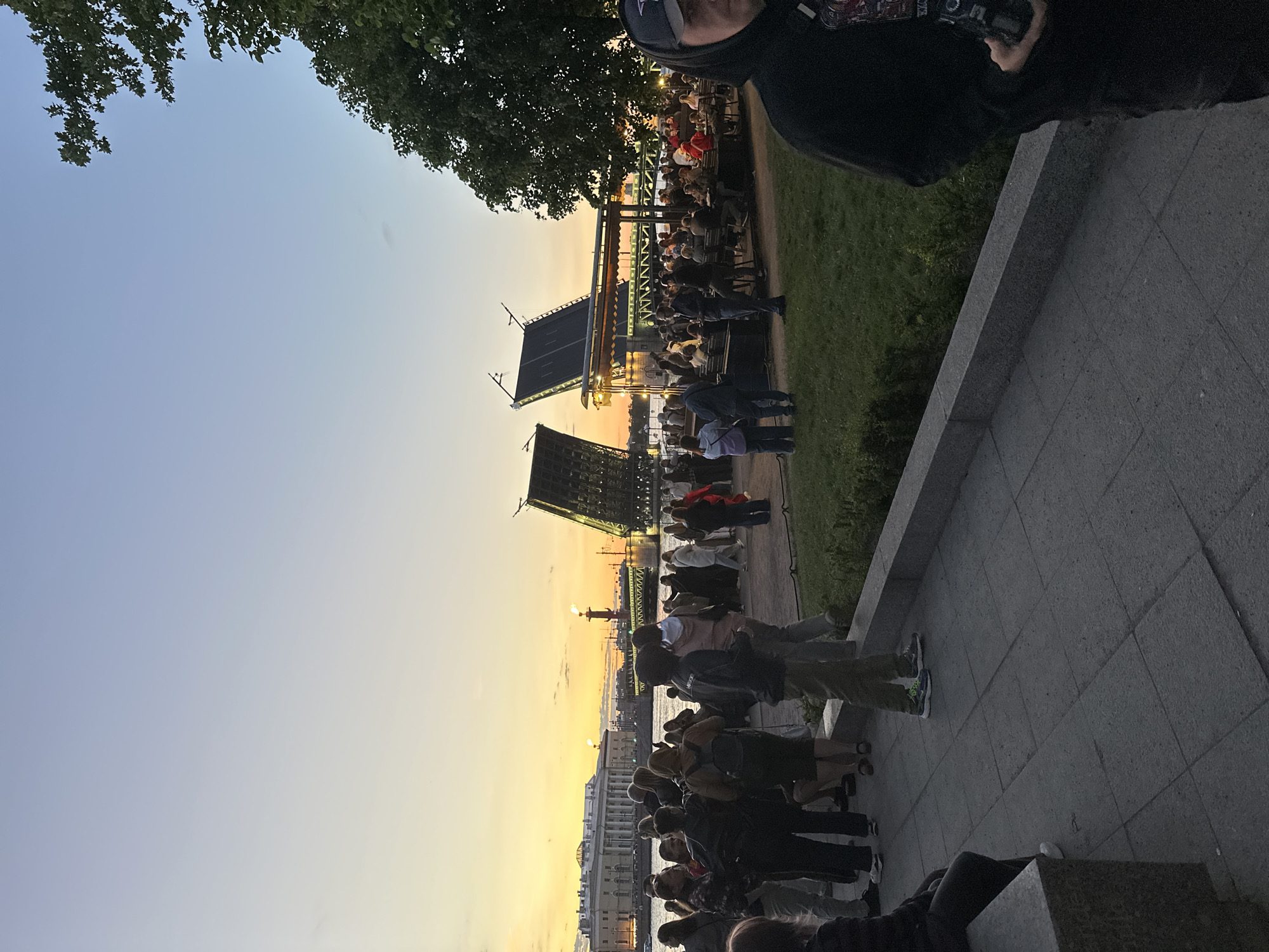 This is a photograph taken in Saint Petersburg, Russia, during sunset. The image captures a large gathering of people on a waterfront promenade, with a drawbridge in the background. The drawbridge is partially raised, revealing its illuminated structure. In the distance, classical buildings with columns are visible, and a tall column with flames on top stands out against the orange sky. People are seen standing and sitting in groups, some appearing to be dining at outdoor tables under a covered area. The foreground shows a paved walkway and a grassy area. The overall atmosphere is one of a public event or gathering by the Neva River during a significant landmark illumination.