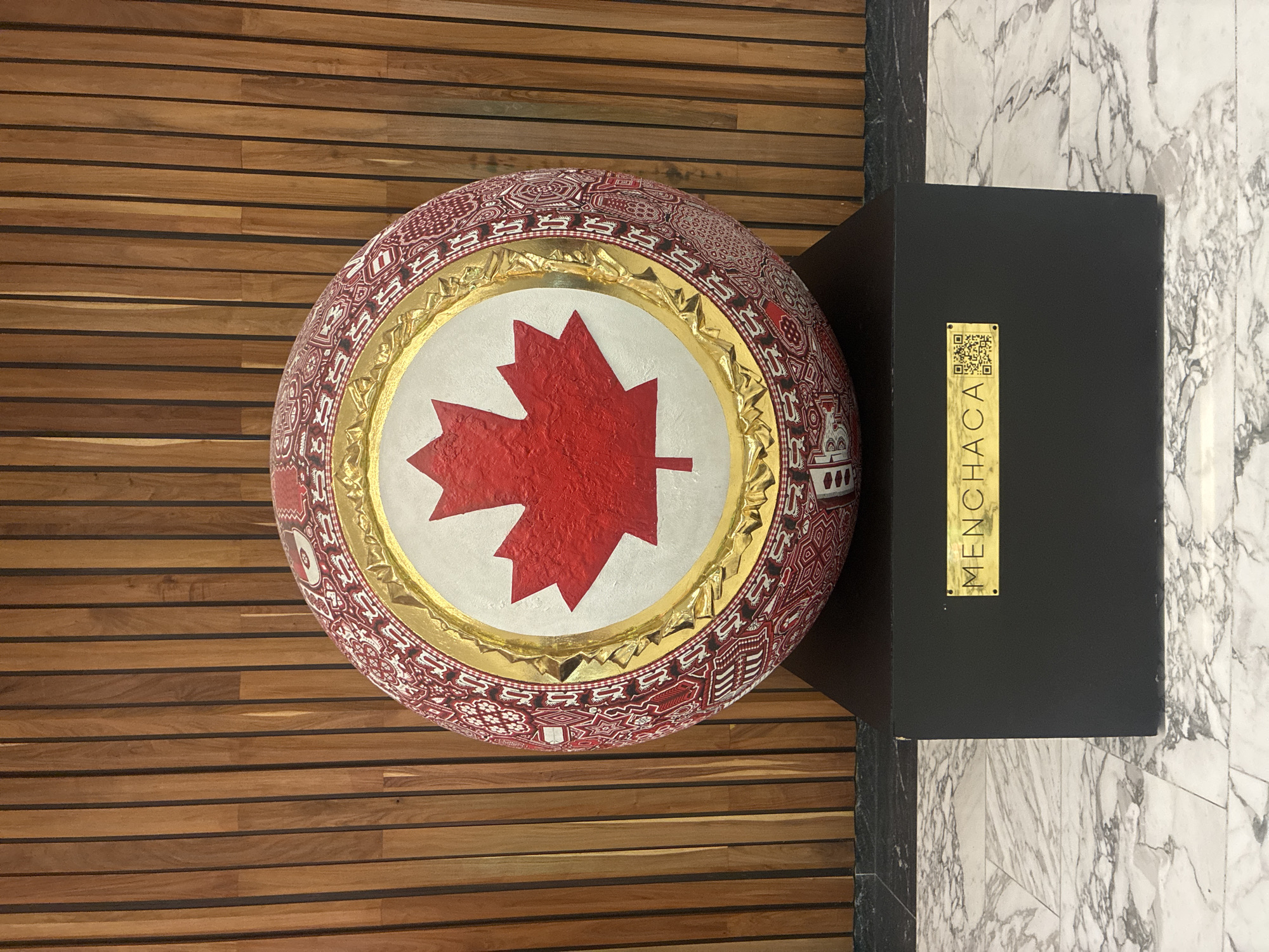 The image shows a spherical sculpture featuring a red maple leaf, the symbol of Canada, centered on a white background surrounded by a golden ring. The sphere's outer surface is adorned with intricate red and white patterns. It rests on a black rectangular pedestal. A gold-colored plaque with the text "MENCHACA" and two QR codes is affixed to the pedestal. The sculpture is located indoors, with wooden paneling visible on the left and white marble flooring on the right. The location of the sculpture is in Mexico City, Mexico.