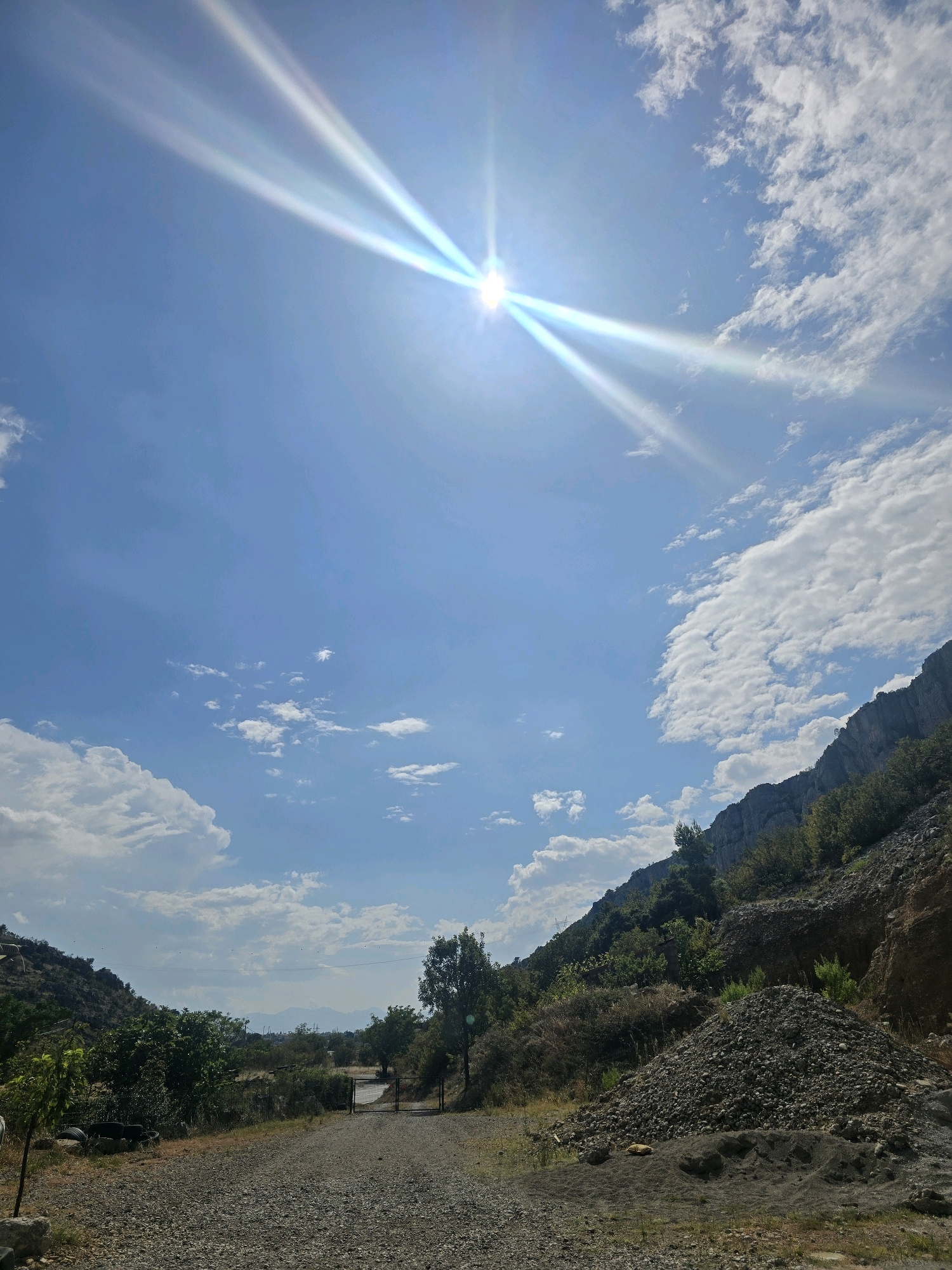 The image depicts a sunny day in a rural area, likely on the outskirts of Podgorica, Montenegro. A dirt road, covered in gravel, leads into the distance, flanked by vegetation and rocky hillsides. The sun is prominently featured in the sky, casting bright rays that create a lens flare effect. Scattered clouds dot the blue sky. The landscape suggests a natural, undeveloped environment with some signs of human activity, such as the road and a metal gate visible in the distance.