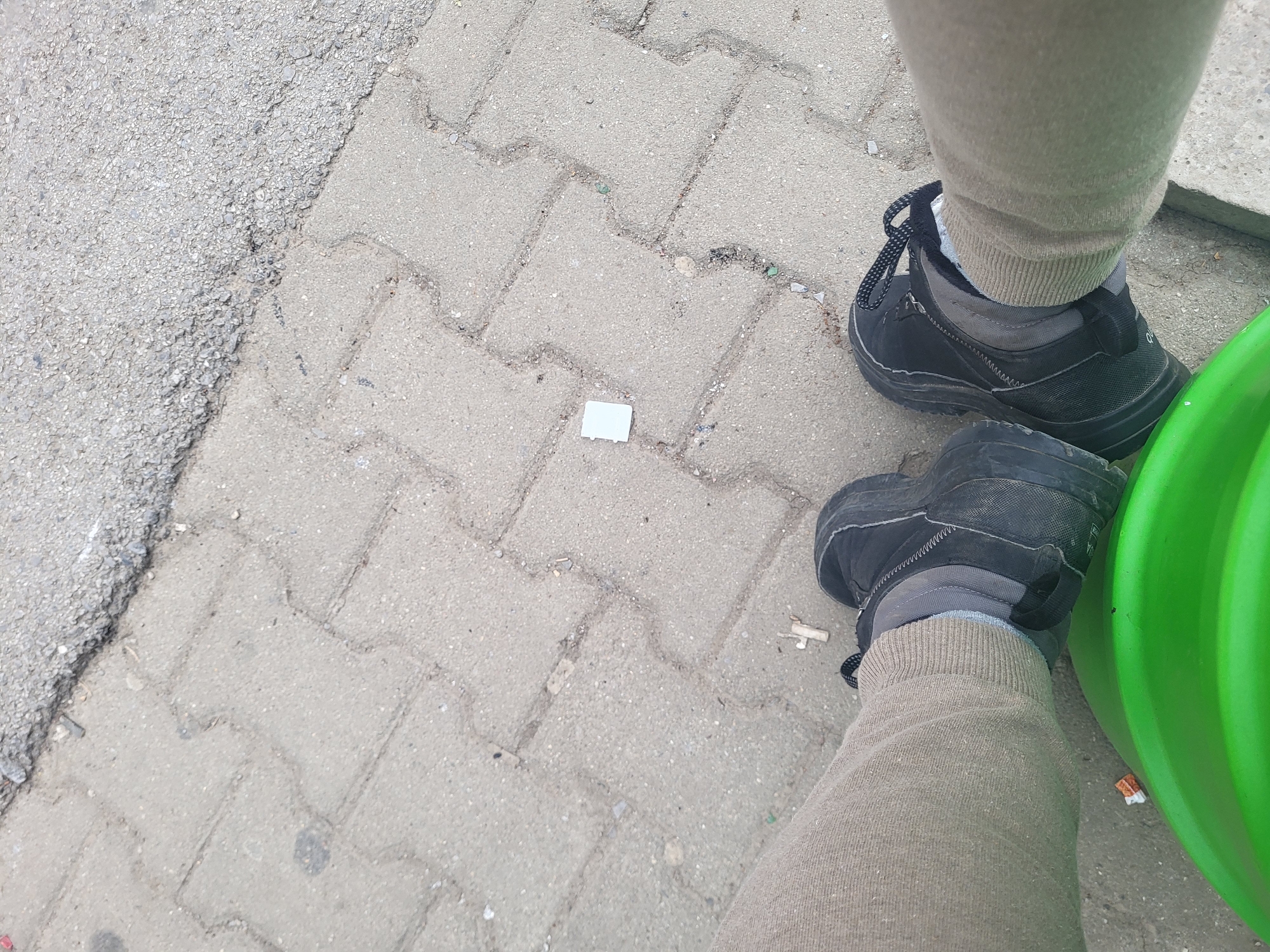 The image shows a street scene in Sofia, Bulgaria. The ground is paved with interlocking concrete blocks and partially bordered by an asphalt edge. Two legs, clad in tan sweatpants and black hiking boots, are visible. One leg is positioned near a bright green plastic object (likely a waste bin). A small white rectangular object lies on the pavement near the center of the frame.