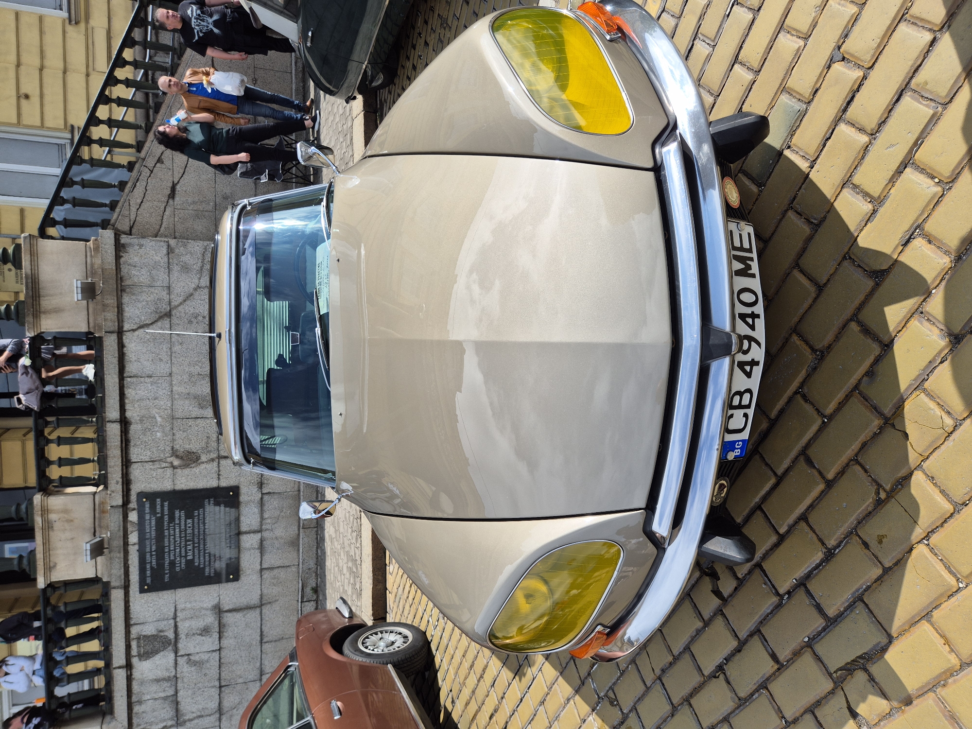 The image shows the front of a vintage light brown car parked on a yellow brick street in Sofia, Bulgaria. The car has distinctive yellow headlights and a Bulgarian license plate that reads "CB 4940 ME".
In the background, a stone building is visible with a black plaque mounted on its wall. The plaque appears to contain text in Bulgarian, possibly commemorating Vasil Levski.
Pedestrians are walking on the sidewalk near the building. They appear to be tourists.
A corner of another car is visible, suggesting there are other vehicles in the vicinity.