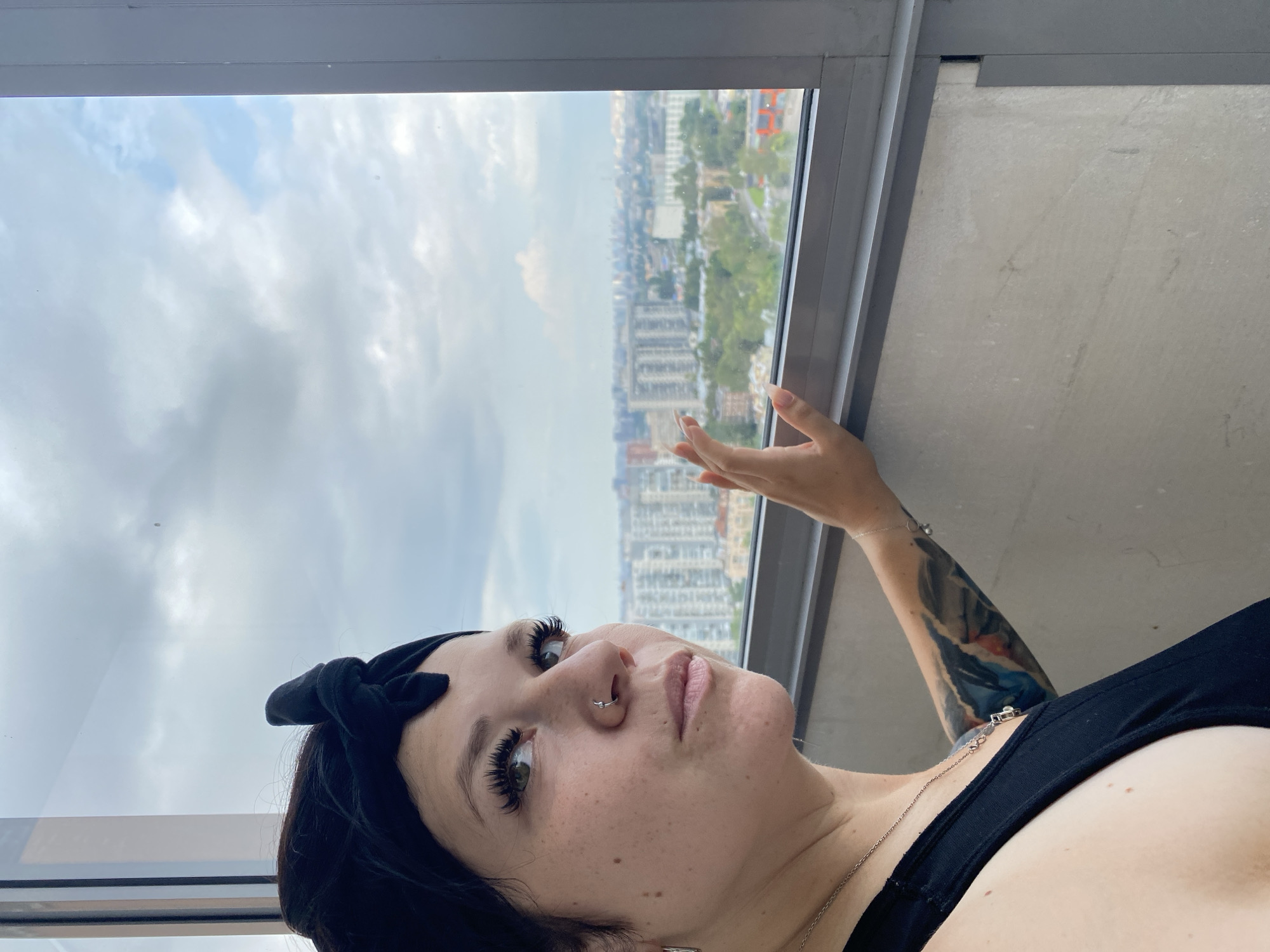 A woman with dark hair and a black headband is looking out a window at a city skyline. She is wearing a black tank top and a necklace. She has tattoos on her arm and a nose ring. The city is visible in the background with buildings and trees. The sky is cloudy. The image is taken from a high vantage point. The woman's hand is resting on the window sill.