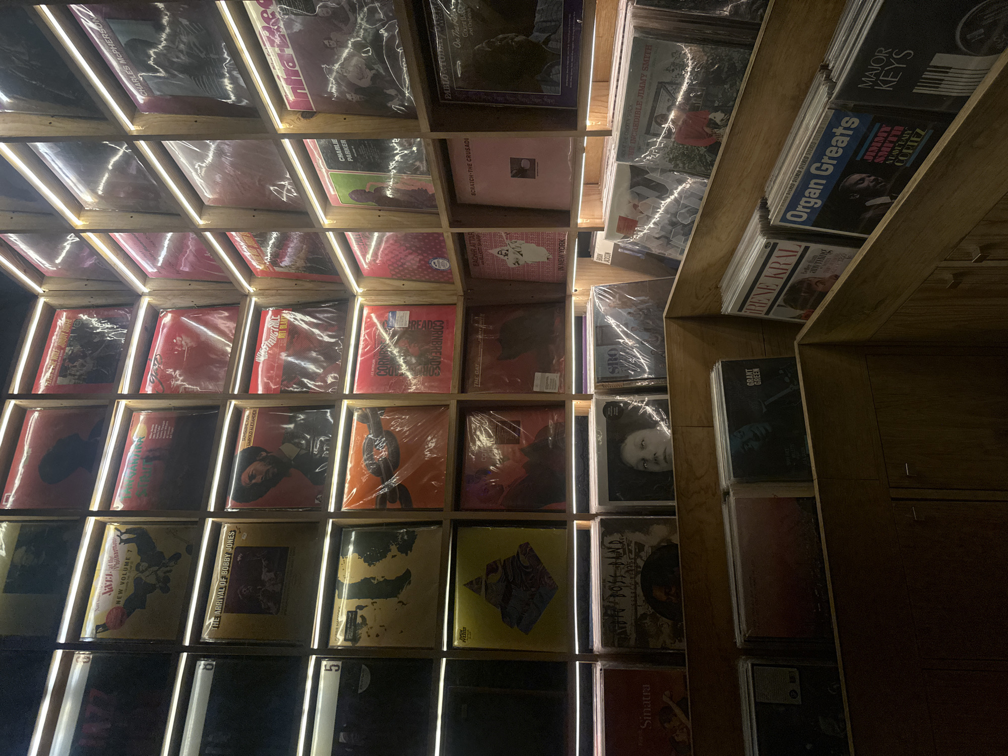 The image shows a tilted view of a wooden shelving unit filled with vinyl records, possibly located in a record store in Mexico City, Mexico. The shelves are illuminated with LED lights, accentuating the triangular patterns of the shelves. The record covers are visible, many of them protected by plastic sleeves. The albums are a variety of styles, including jazz and other genres. Titles such as "Sinatra," "Charlie Parker," "Charles McPherson," "Scratch - The Crusadi," "Pablo Today," and "Organ Greats," along with names like "The Incredible Jimmy Smith" are visible. The wooden structure with the records is against a dark wall.