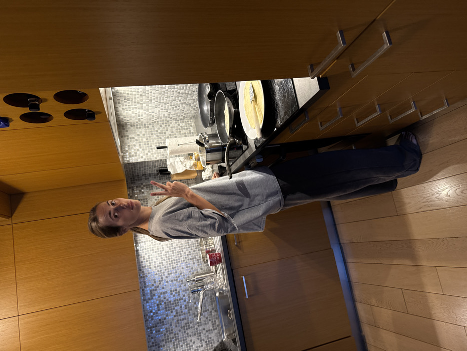 A female individual is present within a modern kitchen setting, identified as being located in Warsaw, Poland. She stands facing the camera, positioned to the right of a kitchen sink and to the left of an electric stovetop. Her body is oriented slightly to the left, and she looks directly at the lens with a neutral expression. Her right hand is raised, displaying the index and middle fingers in a "V" gesture. She has light skin, light brown hair pulled back into a ponytail, and is dressed in an oversized grey t-shirt and dark grey or black wide-leg pants, standing barefoot on a light-toned wooden floor.
The kitchen features light brown, wood-paneled upper and lower cabinetry, with brushed metal handles on the lower cabinets. A reflective, small mosaic tile backsplash extends above dark countertops. A stainless steel sink with a modern faucet and a soap dispenser is visible on the left side of the counter. The stovetop, an electric cooktop, has two non-stick frying pans. One pan contains a round, flat cooking item, consistent with a pancake or crepe. Adjacent to the stovetop, on the dark countertop, a white plate holds a finished pancake or crepe and a yellow plastic spatula. Other objects on the counter include a silver lidded pot, a roll of paper towels on a dispenser, and a partially visible white bag. Warm overhead lighting illuminates the scene, casting soft shadows on the floor.