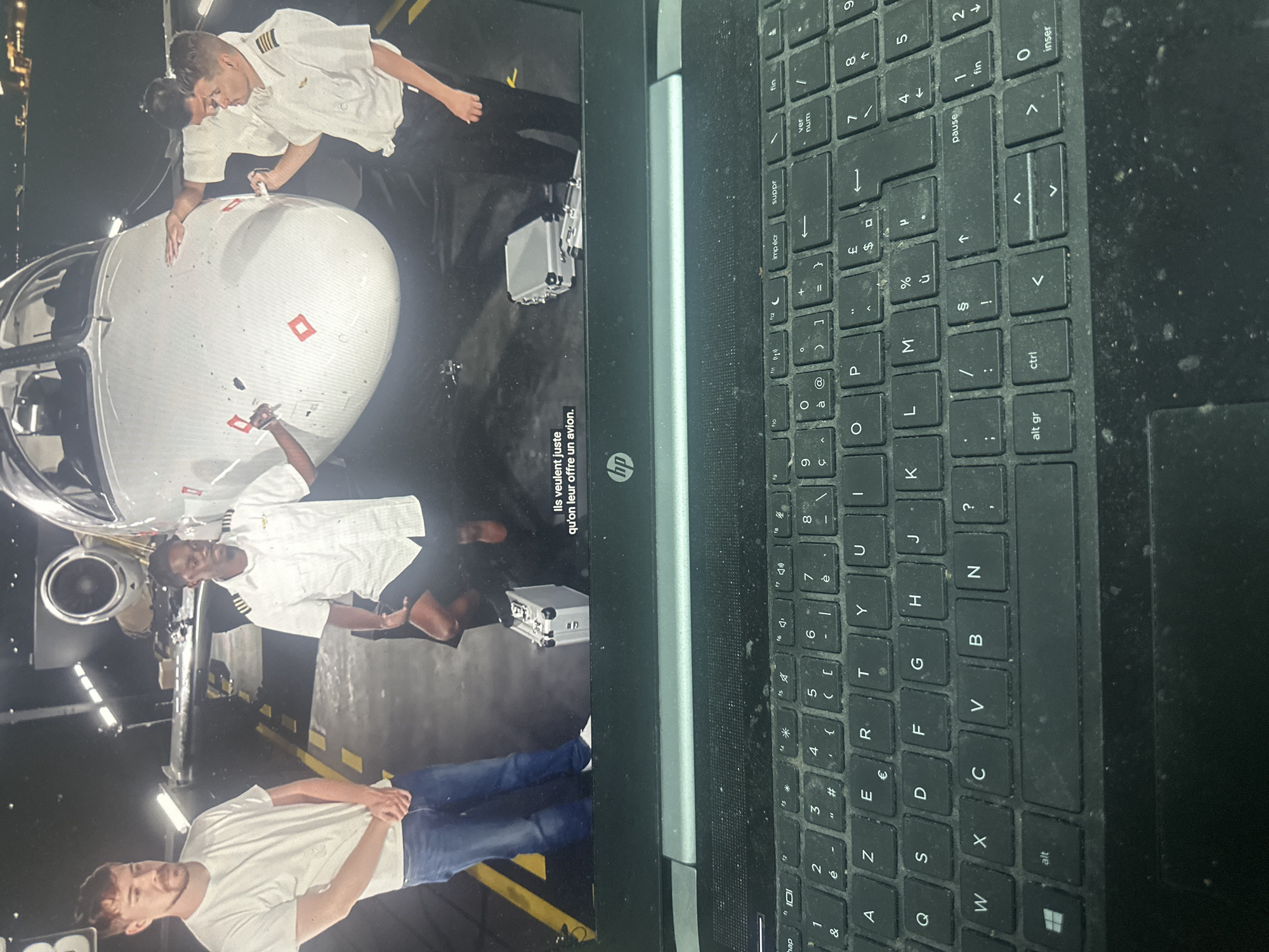 The image displays a laptop computer with its screen showing a video. The laptop's keyboard is visible on the right, appearing dusty and showing a French layout.
The video on the laptop screen depicts four men interacting with a small, white aircraft inside what appears to be an aircraft hangar or maintenance facility in Saint-Leu-d'Esserent, France. The setting is dimly lit, with artificial overhead lights illuminating the scene.
Two of the men are wearing pilot uniforms with white shirts and epaulets. One pilot is leaning over the aircraft's nose cone, seemingly inspecting it or explaining something. Another man in a white t-shirt is also close to the nose cone, looking at it. A second pilot stands further back near the aircraft's engine, which is a turbofan. The fourth man, wearing a white t-shirt and blue jeans, stands further away, observing the activity. A silver briefcase and another similar case are visible on the floor near the aircraft.
The visible text on the laptop screen is in French: "Ils veulent juste qu'on leur offre un avion," which translates to "They just want to be offered a plane." The laptop itself is an HP model, indicated by the "hp" logo. The keyboard features keys such as "alt gr", "ctrl", "inser", "suppr", "imp écr", "pause", and "ver num", along with French-specific characters.
