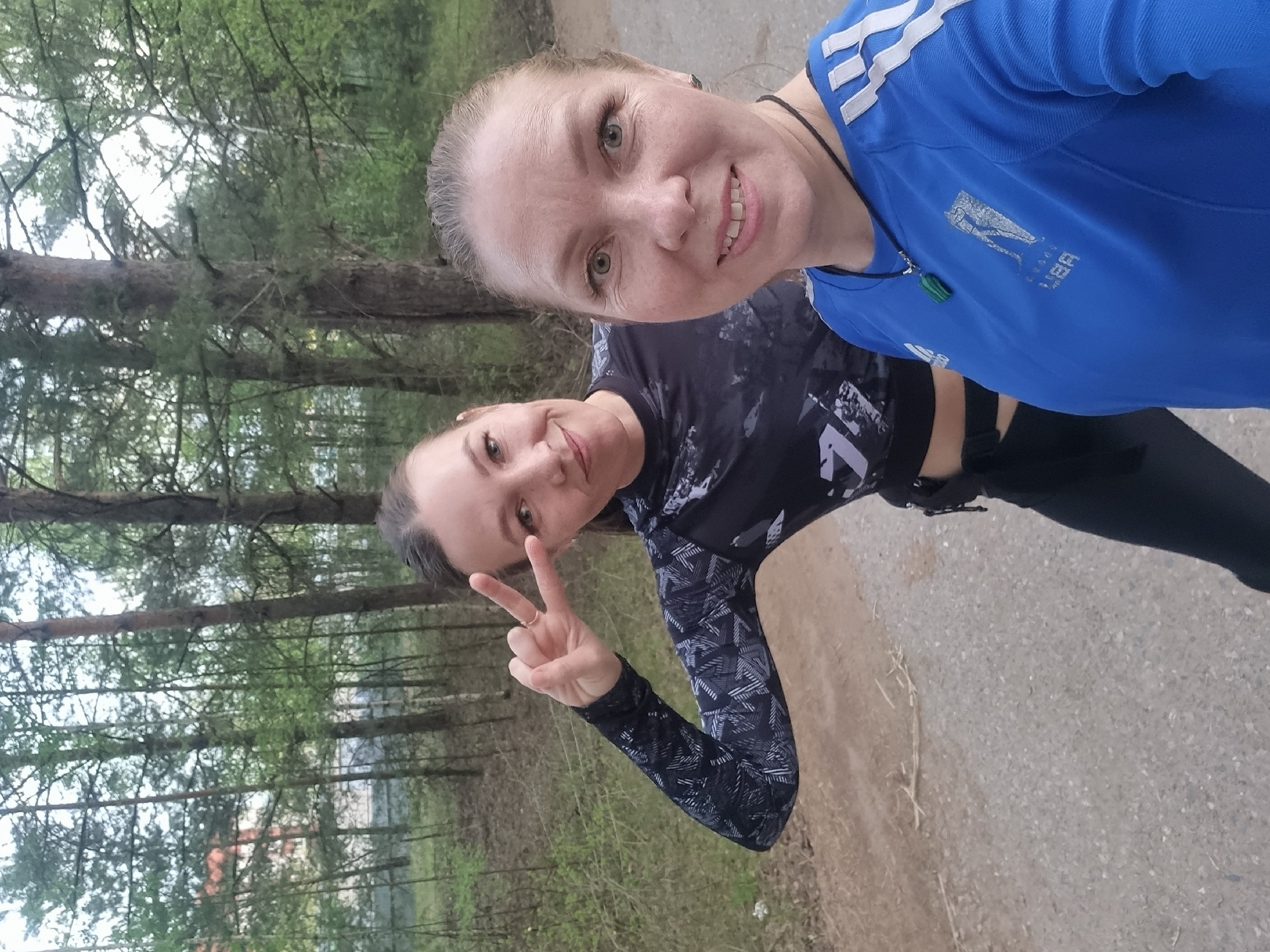 Two women pose for a selfie on what appears to be an asphalt road, possibly a walking or jogging path in a wooded area. The woman in the foreground wears a long-sleeved, dark athletic shirt with a geometric pattern and is holding up a peace sign. The woman behind her is wearing a blue athletic shirt with white stripes on the sleeves and a pendant necklace. The background consists of trees and green foliage. Based on the visual context, it's probable that this image was taken in Chaikovsky, Russia, as requested.