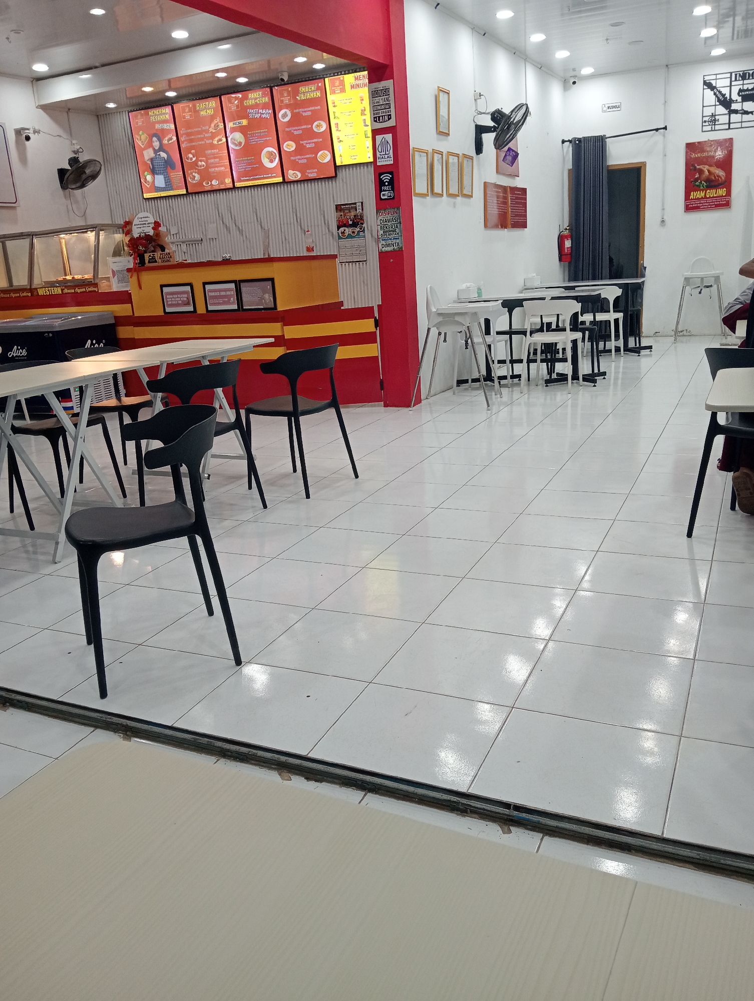 This image depicts the interior of a casual dining establishment, likely a fast-food restaurant, in Pringsewu, Indonesia. The restaurant features white tiled flooring, bright fluorescent lighting, and white walls. A prominent red-and-yellow counter area is visible, displaying menus and other restaurant information. Several tables and chairs, including black plastic chairs and white high-top chairs, are arranged throughout the dining space. In the background, there is a promotional poster for "Ayam Guling" (roast chicken) and a sign indicating "MUSHOLLA" (prayer room). The overall impression is a clean and functional dining environment typical of a local eatery.