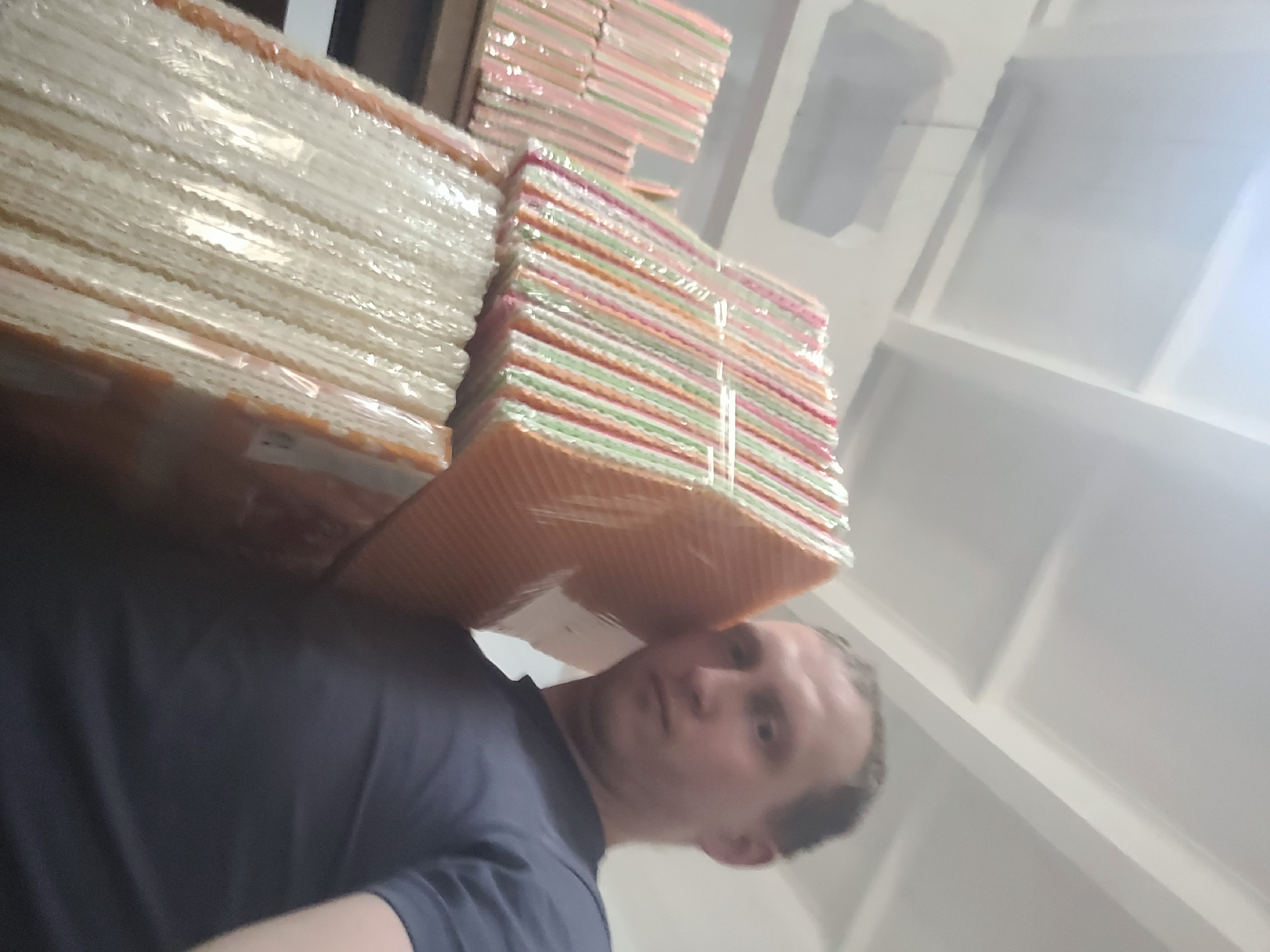 A man stands in a dimly lit warehouse, holding a stack of colorful, individually wrapped wafer cookies. The cookies are in various shades of pink, orange, and green. The man is wearing a dark blue t-shirt. The warehouse ceiling has visible beams and lighting fixtures. The floor is not visible.