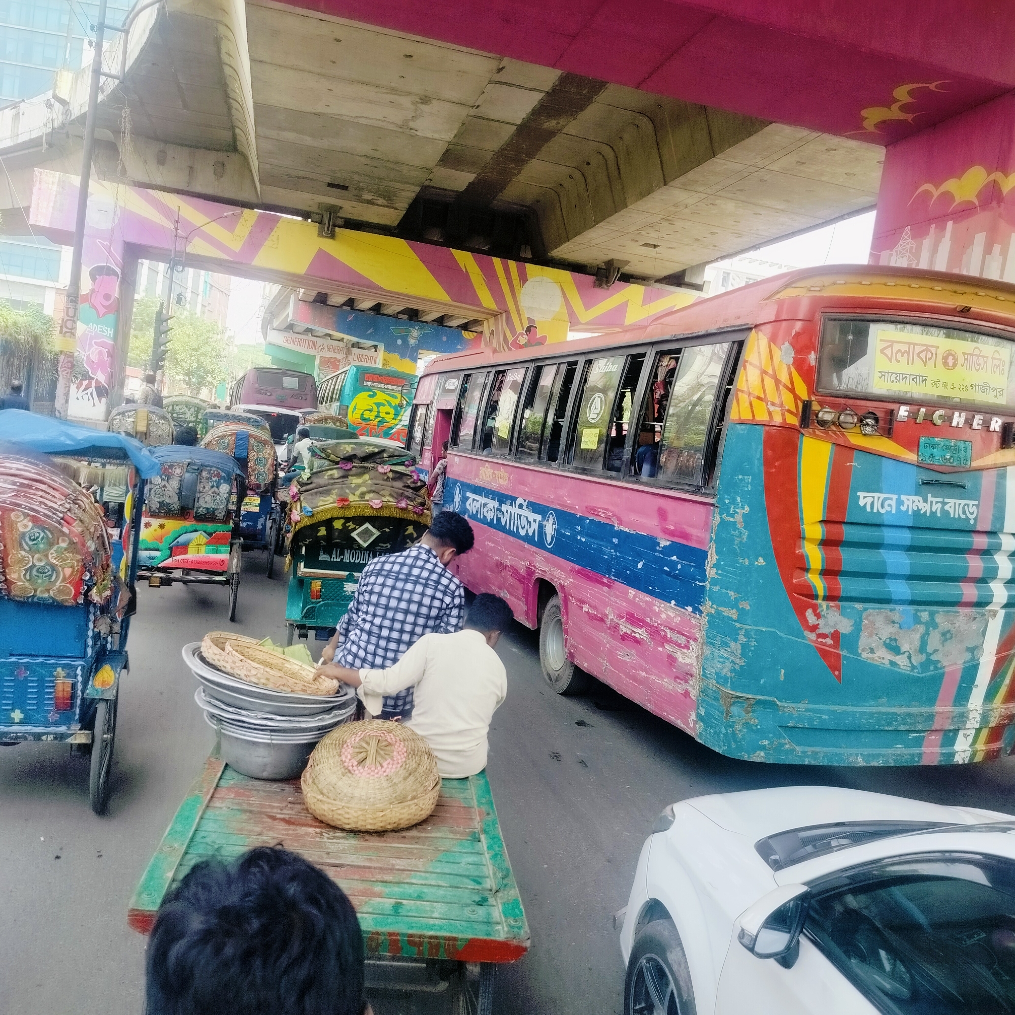 The image depicts a busy street in Dhaka, Bangladesh, filled with various forms of transportation, including rickshaws, a bus, and a white car. Two men are seen near a cart loaded with baskets and metal bowls, possibly engaged in trade or transport of goods. The bus, labeled "Balaka Service Ltd," indicates a local transportation service. Overhead, a brightly painted overpass adds color to the urban scene. The presence of an EICHER bus and the signage "Sayedabad Route No. A-225 Gazipur" pinpoints the location within Dhaka. There are rickshaws in the street as well.