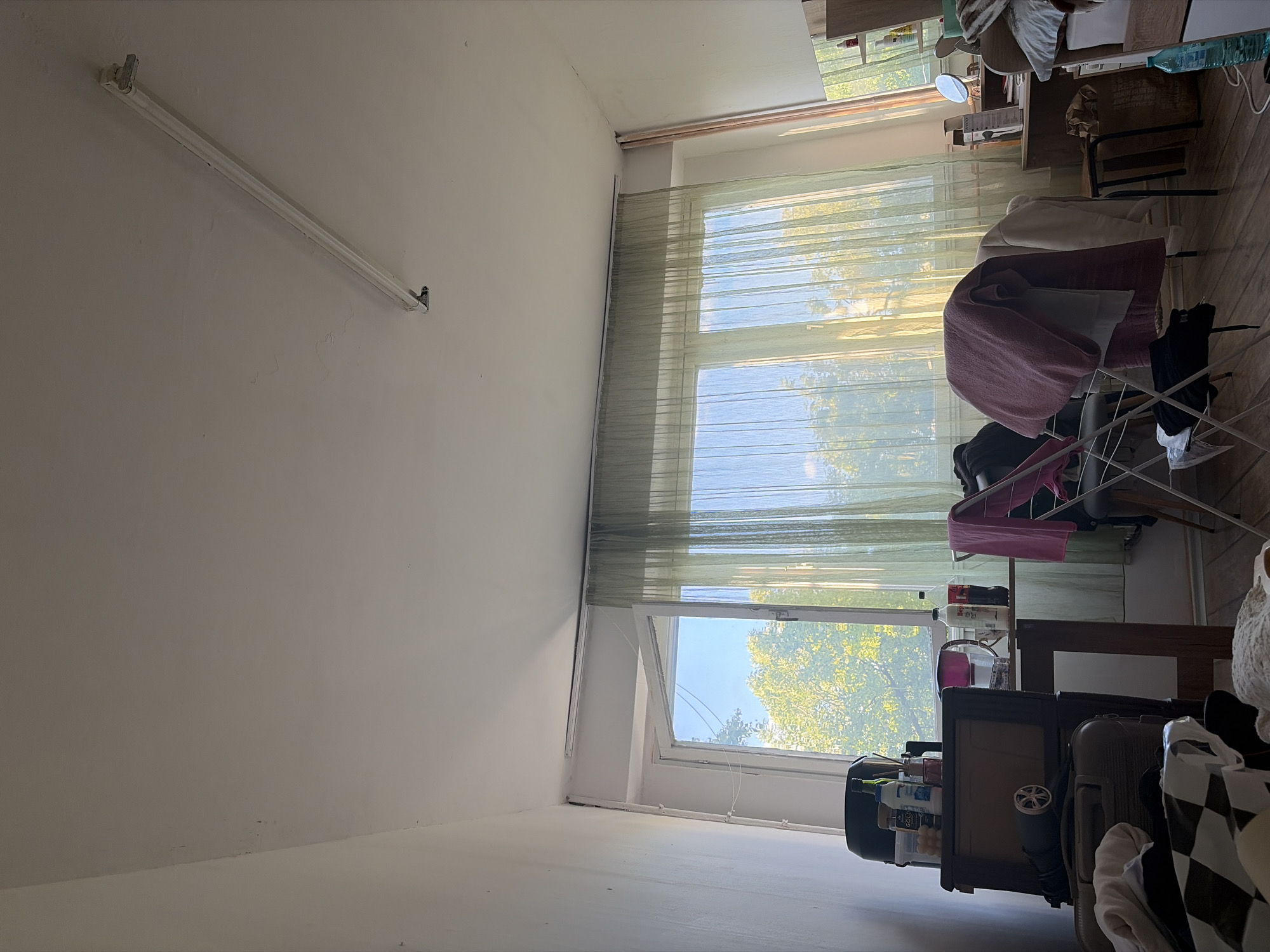 The image shows an interior space. A white, fluorescent tube light fixture is mounted horizontally on a white wall in the upper left of the frame. A long, sheer, green curtain covers a window, which is partly visible. Through the window, trees and blue sky are visible. In front of the window, a room setting includes a desk with miscellaneous objects. A clothing rack is partially covered with a pink towel and black clothing. Other objects are indistinct due to the angle and proximity. It's impossible to determine the location in Sofia, Bulgaria, without further information.