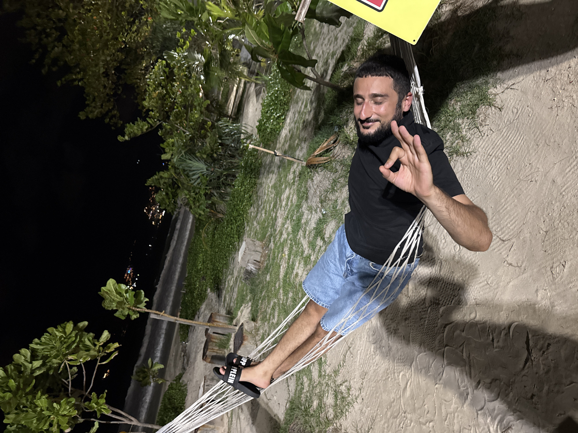 A man with dark hair and a beard sits in a hammock on a beach in Patong, Thailand. He wears a black t-shirt and denim shorts, and black sandals with white text. The hammock is strung between trees. The man is smiling and giving an "OK" hand gesture. Behind him, there is sand, vegetation, and glimpses of the city lights at night.