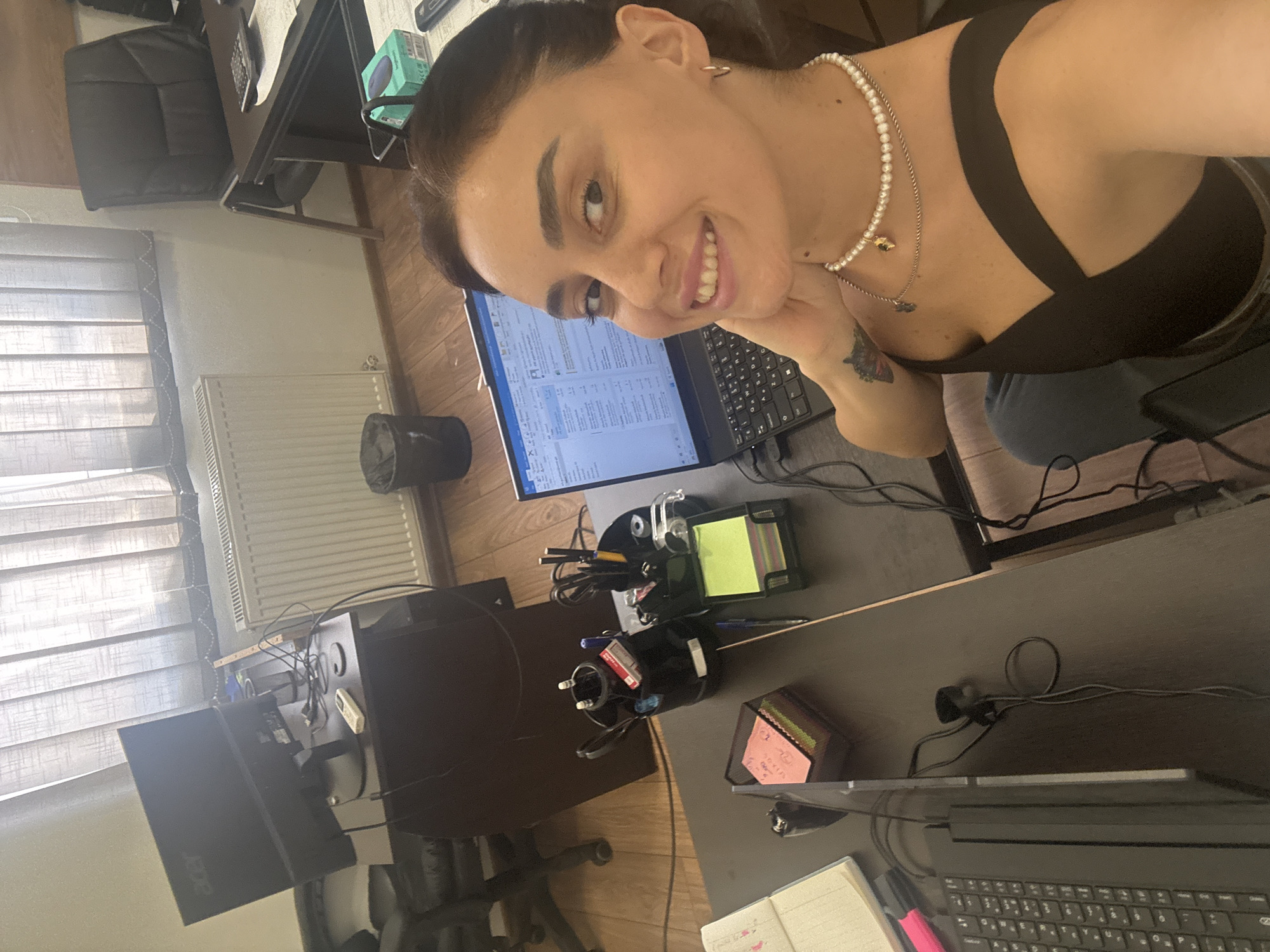 A woman is taking a selfie in what appears to be an office setting. She is smiling and looking at the camera, with her hand resting on her chin. She is wearing a black top and a pearl necklace. There is a laptop in front of her, and a desk with various office supplies, including pens, sticky notes, and a notebook. In the background, there is a computer monitor, a radiator, and blinds on the window. The lighting suggests it is daytime. The specific location in Tbilisi, Georgia, is not discernible from the image.