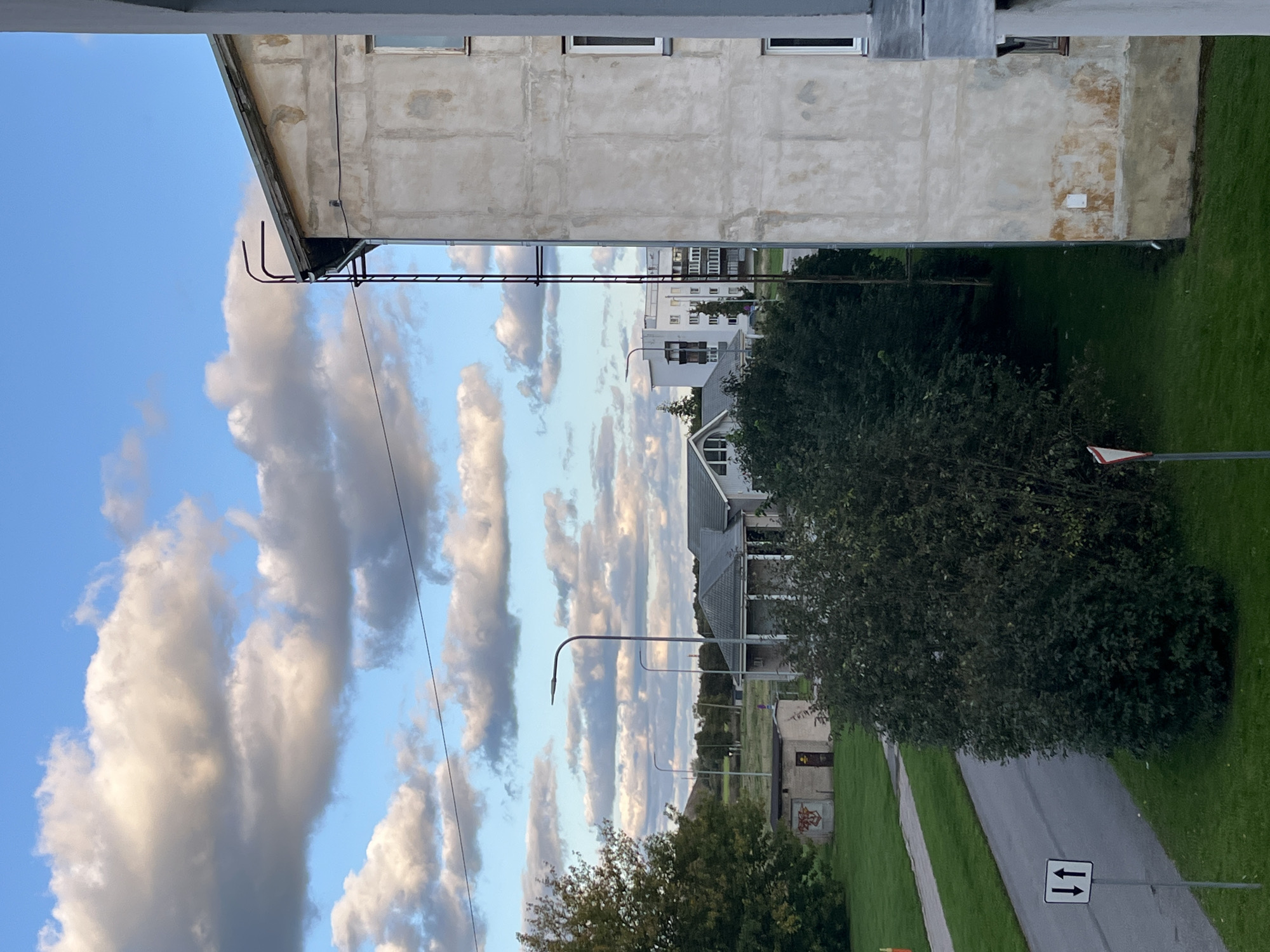 The image shows an outdoor scene with a building in the foreground, an apartment building to the left, and a street. The sky is partly cloudy with white and gray clouds against a blue sky. A tall, dark ladder is attached to the side of the building in the foreground, reaching towards the roof. There are windows on the building's facade. To the left of the building, a paved road curves through a grassy area. Several streetlights are visible along the road. Further in the background, there are more buildings, including what appears to be a house and a multi-story apartment building. Trees and bushes are scattered throughout the landscape. A traffic sign with two vertical arrows, one pointing up and one pointing down, is located on the side of the road. The overall impression is of a residential or urban area in Estonia.