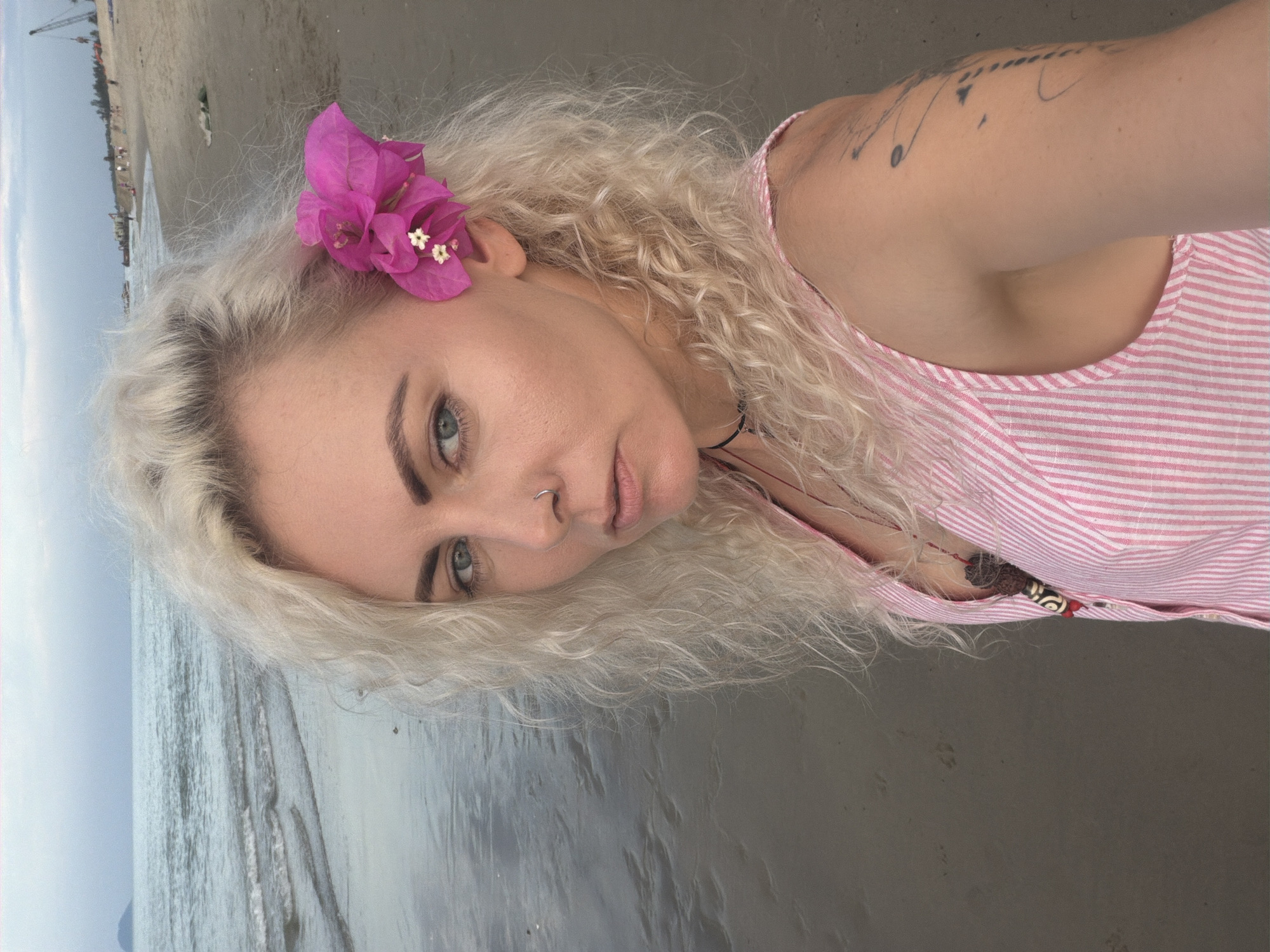 A woman with blonde, curly hair is taking a selfie on a beach in Hoi An, Vietnam. She is wearing a pink and white striped tank top and has a pink flower tucked behind her ear. Her eyes are blue and she is wearing a nose ring. The ocean is visible behind her, with gentle waves lapping at the shore. The sky is overcast. In the background, there are people and construction equipment on the beach. The sand is wet and reflects the sky.