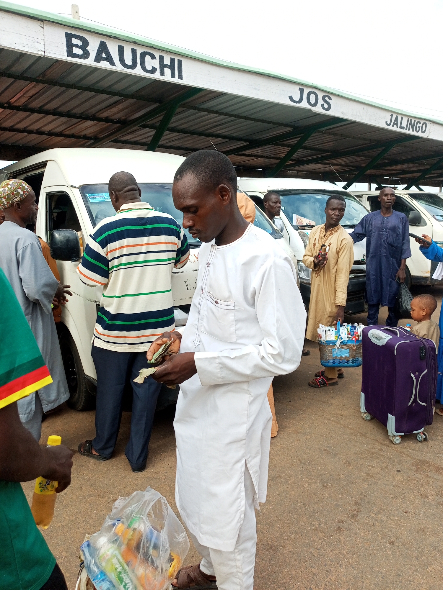 The image depicts an outdoor scene at what appears to be a bus station or transportation hub, indicated by the presence of several minibuses and overhead signage. The signs above read "BAUCHI," "JOS," and "JALINGO," suggesting these are destinations or origins for the transport.
In the foreground, a man in a white traditional outfit is holding cash and what appears to be dried leaves or plant matter in his hands, looking down at them. Another person in a green shirt with yellow and red trim is holding a yellow bottle of drink. To the left, a man wearing a patterned cap and a grey robe is interacting with someone inside a white minibus.
Behind the foreground individuals, more people are visible. One man wears a striped polo shirt and dark pants. Another man, in a beige outfit, carries a basket filled with small boxes, possibly medication or toiletries, and is holding a few bottles. A man in a dark blue outfit stands further back. A young child in a beige outfit is also present near a large purple suitcase with wheels.
The ground is paved, and the lighting suggests it is daytime. The overall atmosphere seems to be one of travel and commerce.