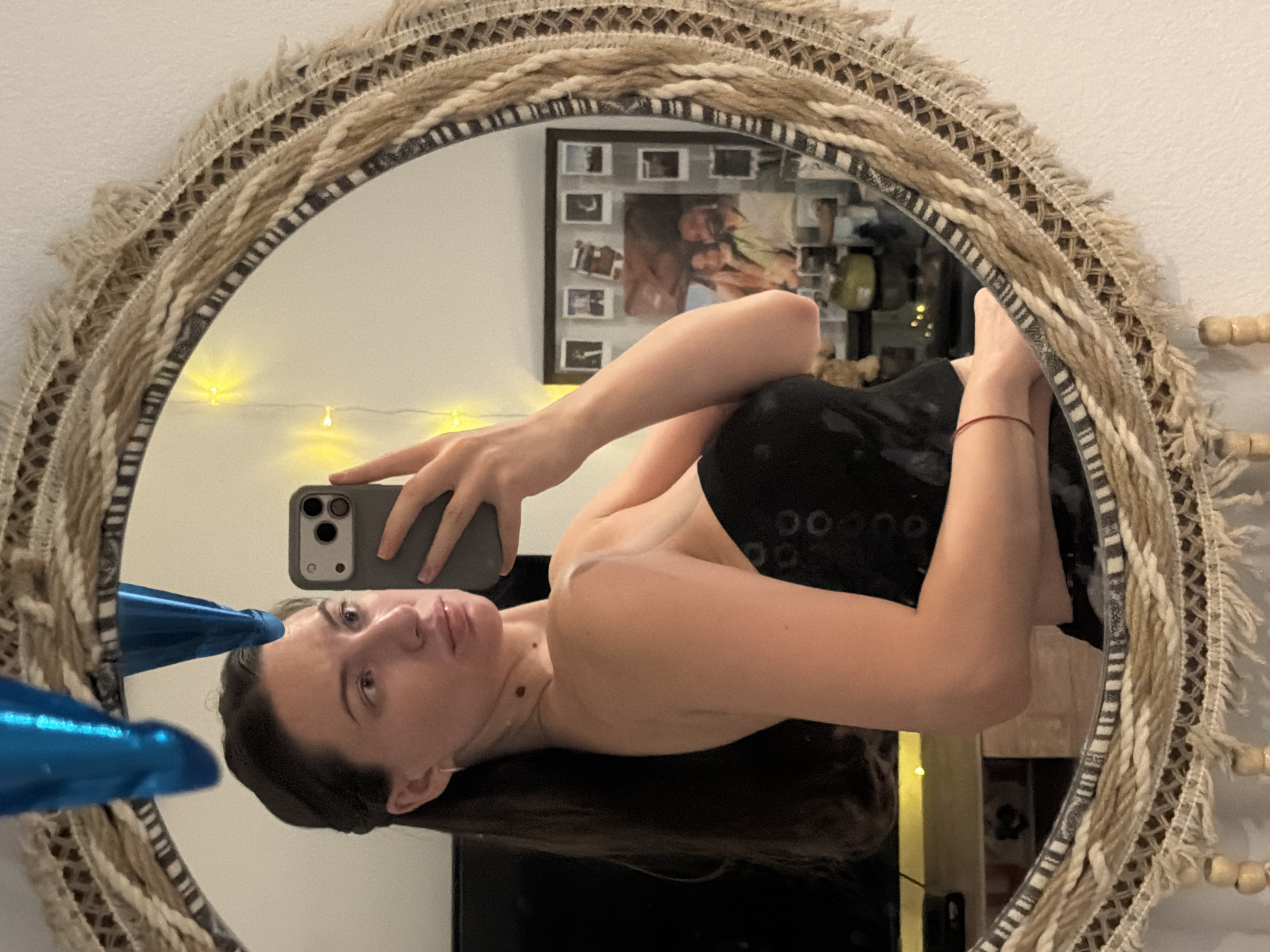 A woman is taking a selfie in a circular mirror. The mirror has a textured, woven frame with fringe. The woman has long, dark hair and is wearing a black strapless top. She is holding a smartphone in her right hand. In the reflection of the mirror, a framed collage of photographs is visible on the wall behind her, along with some yellow fairy lights and a blue balloon. A television is also partially visible in the reflection. The setting appears to be an indoor environment, possibly a bedroom or living area. The image does not contain any specific visual cues to identify its location as Sharm El Sheikh, Egypt.