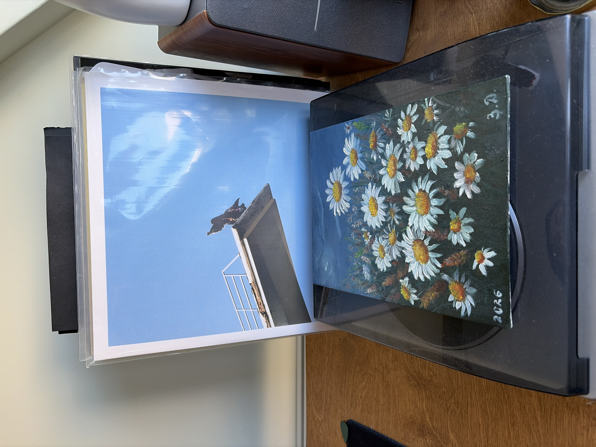 In an indoor setting, a vinyl record player is positioned on a wooden surface. On the record player's platter rests a small, framed painting depicting a field of white daisies with yellow centers against a dark background. To the left of the painting, the number "2026" is visible, and to the right, the initials "3.D." are painted. Behind the painting, an album cover featuring a bird perched on a diving board under a blue sky is leaning against a black mat. A portion of a wooden speaker is visible to the right of the album cover. The overall lighting suggests an indoor, daytime environment.