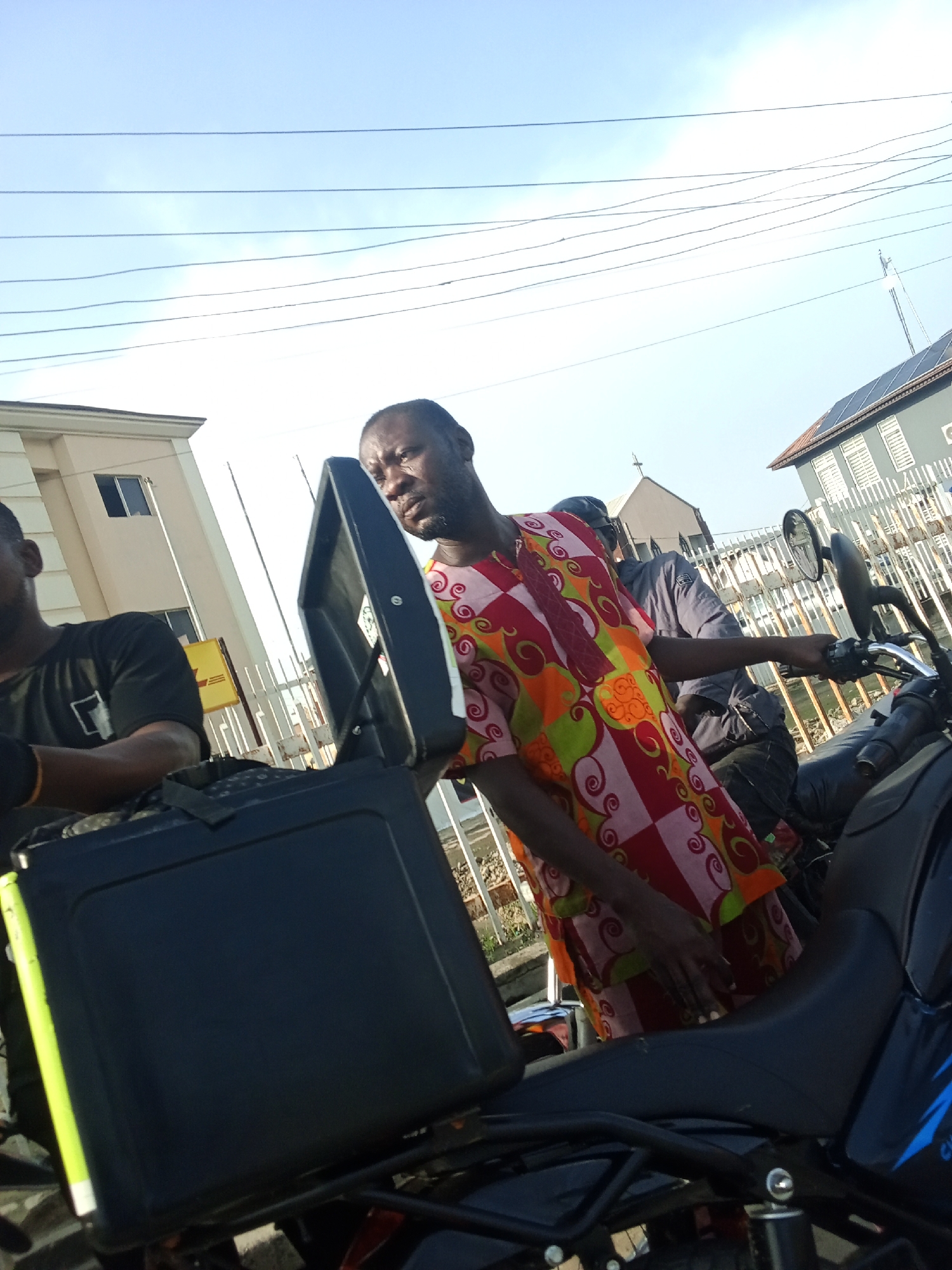 The image depicts two men on motorcycles in what appears to be the city of Ikorodu, Nigeria. One man, closest to the camera, is wearing a black t-shirt. The other man stands next to a motorcycle, wearing a colorful patterned shirt. A black carrier box is mounted on one of the motorcycles. Buildings and power lines are visible in the background, along with another person partially visible behind the man in the colorful shirt.