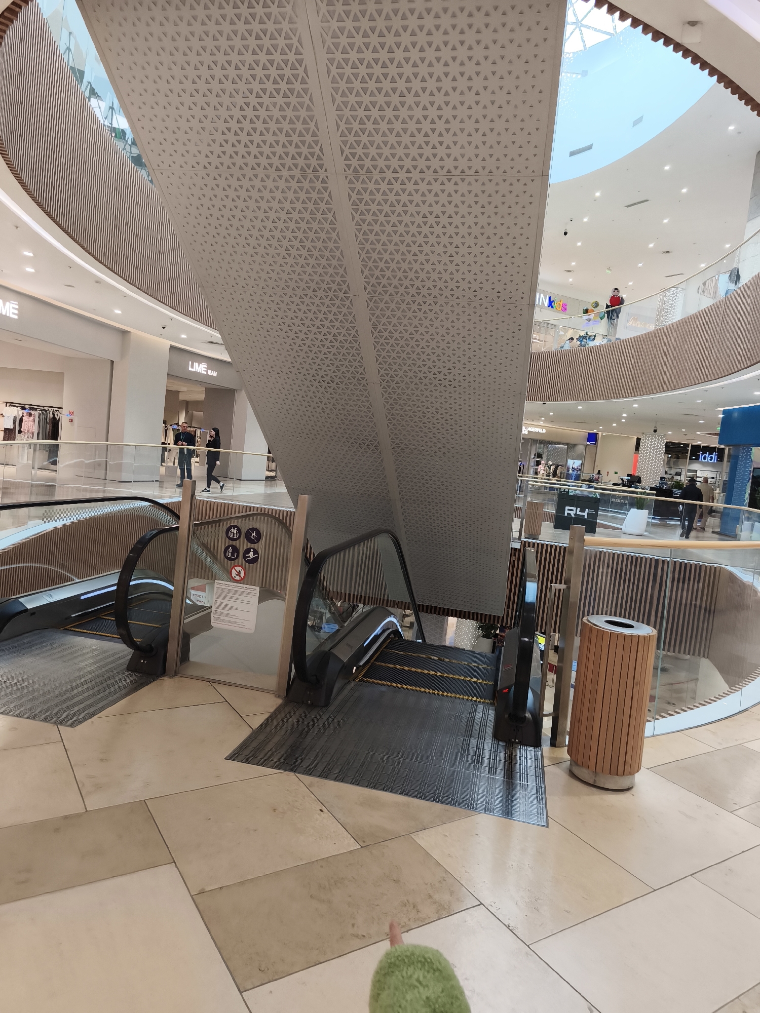 The image depicts an interior view of a multi-level shopping mall, likely in Vladivostok, Russia, given the presence of retail establishments like "LIME MAN" and "INKIDS" which are common brands in the region. Two escalators, one ascending and one descending, are prominently featured in the foreground. The escalators have black treads with yellow safety lines and are enclosed by clear glass railings and wooden paneling. A cylindrical wooden trash receptacle stands to the right of the descending escalator.
In the mid-ground, people can be seen walking on the second level of the mall, browsing in shops. One individual in a red shirt is standing near the railing, looking down towards the escalators. Retail storefronts with illuminated signage are visible, including "LIME MAN," "INKIDS," "R4," and "ididi." The mall's architecture is modern, with curved balconies, extensive use of wood paneling, and a large, geometric patterned ceiling that extends upwards towards a skylight. The floor is tiled with large, square beige and light brown tiles.
In the lower right corner, a green, fuzzy object, possibly a toy or a piece of clothing, is visible, with a finger extending from it and pointing towards the escalators. The overall environment suggests a bright, busy commercial space.