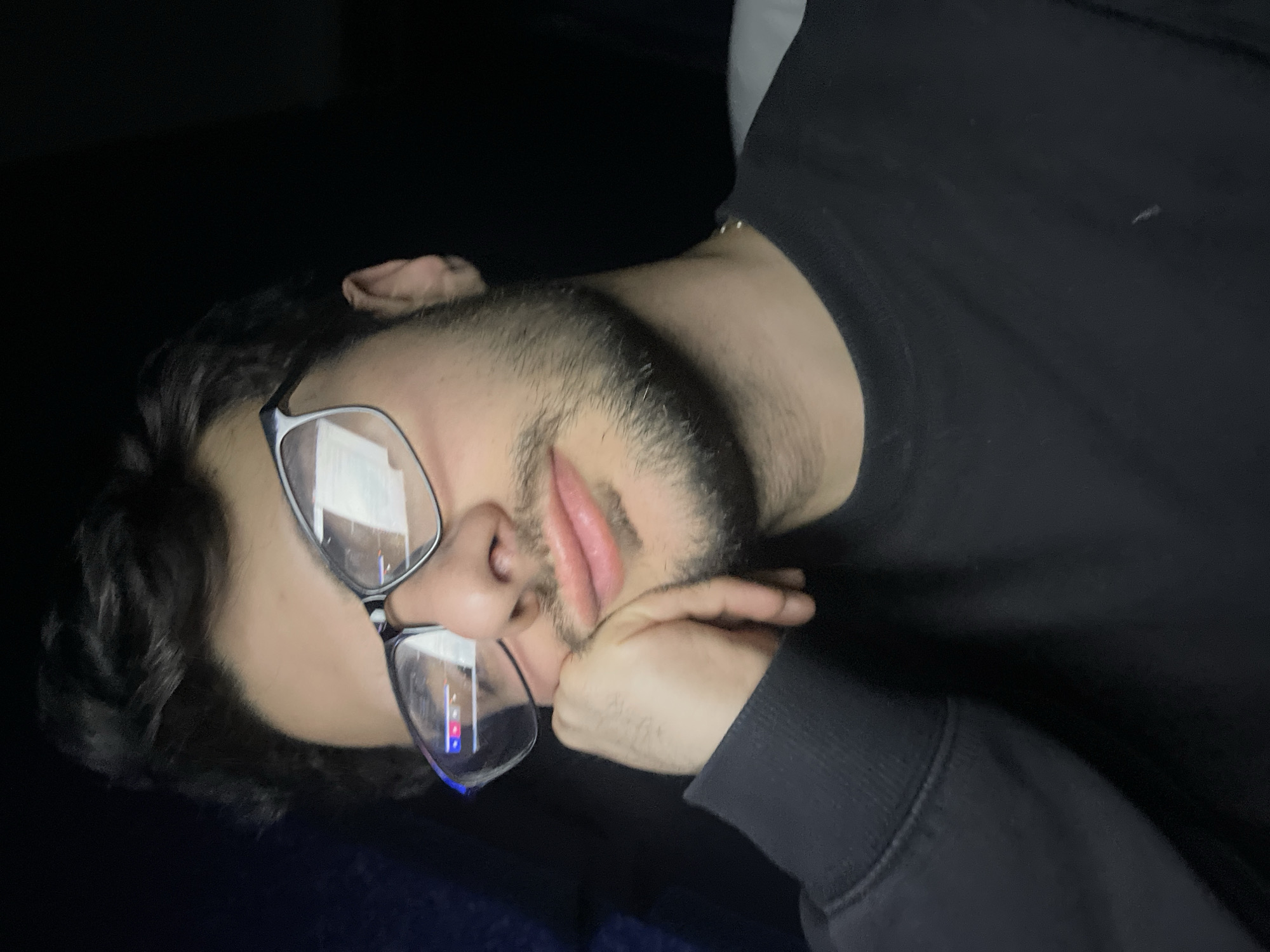 A person is lying down, supporting their head with a hand. They are wearing glasses and a dark-colored shirt. The background is mostly dark, suggesting a dimly lit room. The image does not contain any information to determine a location in Tbilisi, Georgia.