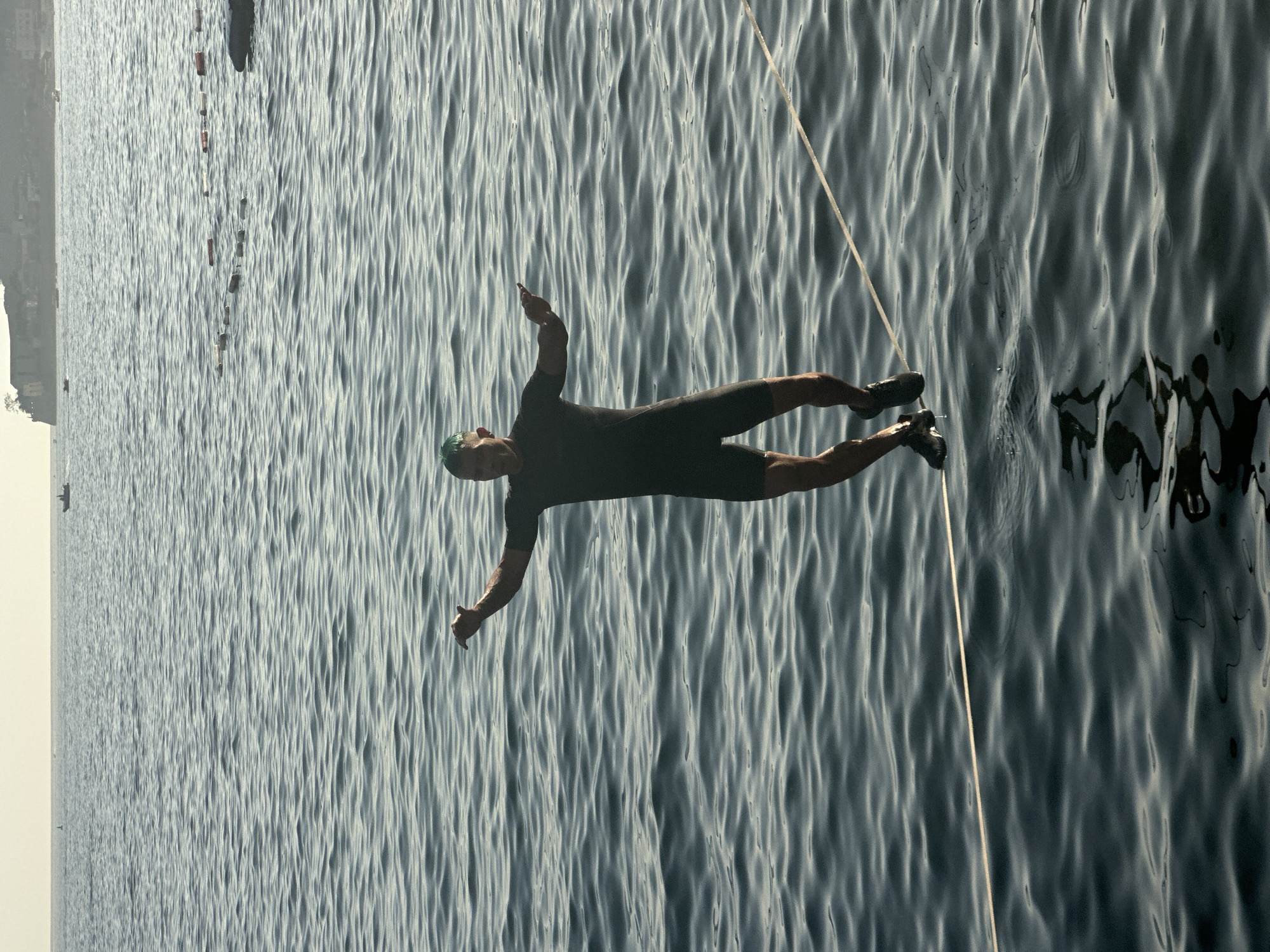 A man is slacklining across the water in Budva, Montenegro. He is wearing a dark, short-sleeved wetsuit and black shoes. His arms are extended for balance, and he appears to be mid-stride. The slackline is a light-colored rope, taut and stretched across the choppy, dark blue water. In the background, the hazy silhouette of the Budva coastline is visible, with buildings perched on a hillside. Several buoys mark a section of the water further out, and a small boat can be seen in the distance. The sky is a pale, overcast hue, suggesting either early morning or late afternoon. The overall scene captures a moment of extreme balance and athleticism against a coastal backdrop.