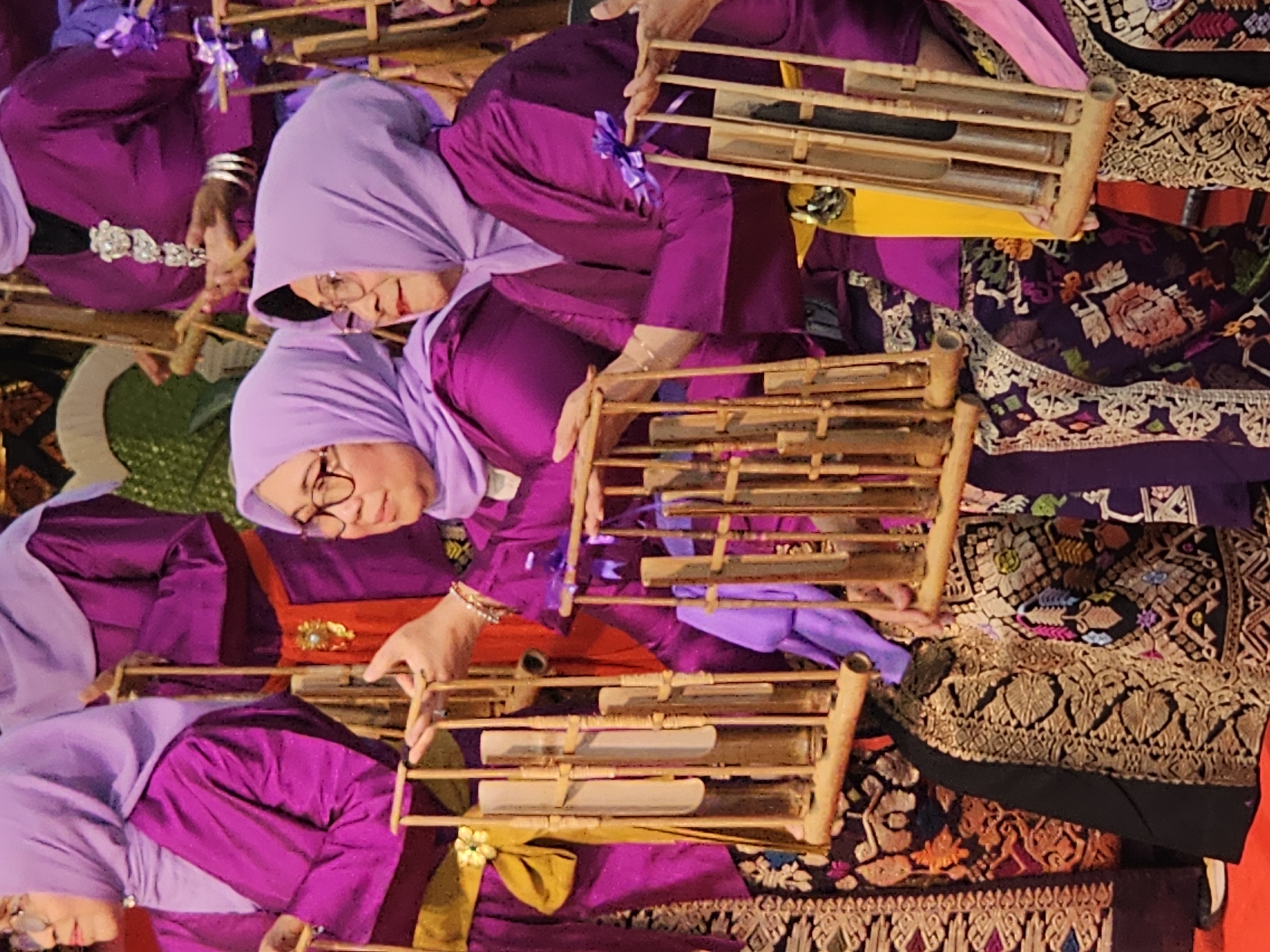 The image captures a group of women in traditional purple attire and headscarves, holding musical instruments made of bamboo. These instruments, known as Angklung, are arranged in vertical racks with multiple tubes. The women are positioned in what appears to be an indoor setting, possibly a performance space or a cultural event. The women are all wearing glasses, and their hands are visible holding the instruments, suggesting they are either playing or preparing to play. The background is out of focus, but hints of ornate fabric and decorative elements are visible, indicating a formal or ceremonial occasion. The overall impression is one of cultural performance and artistic expression.