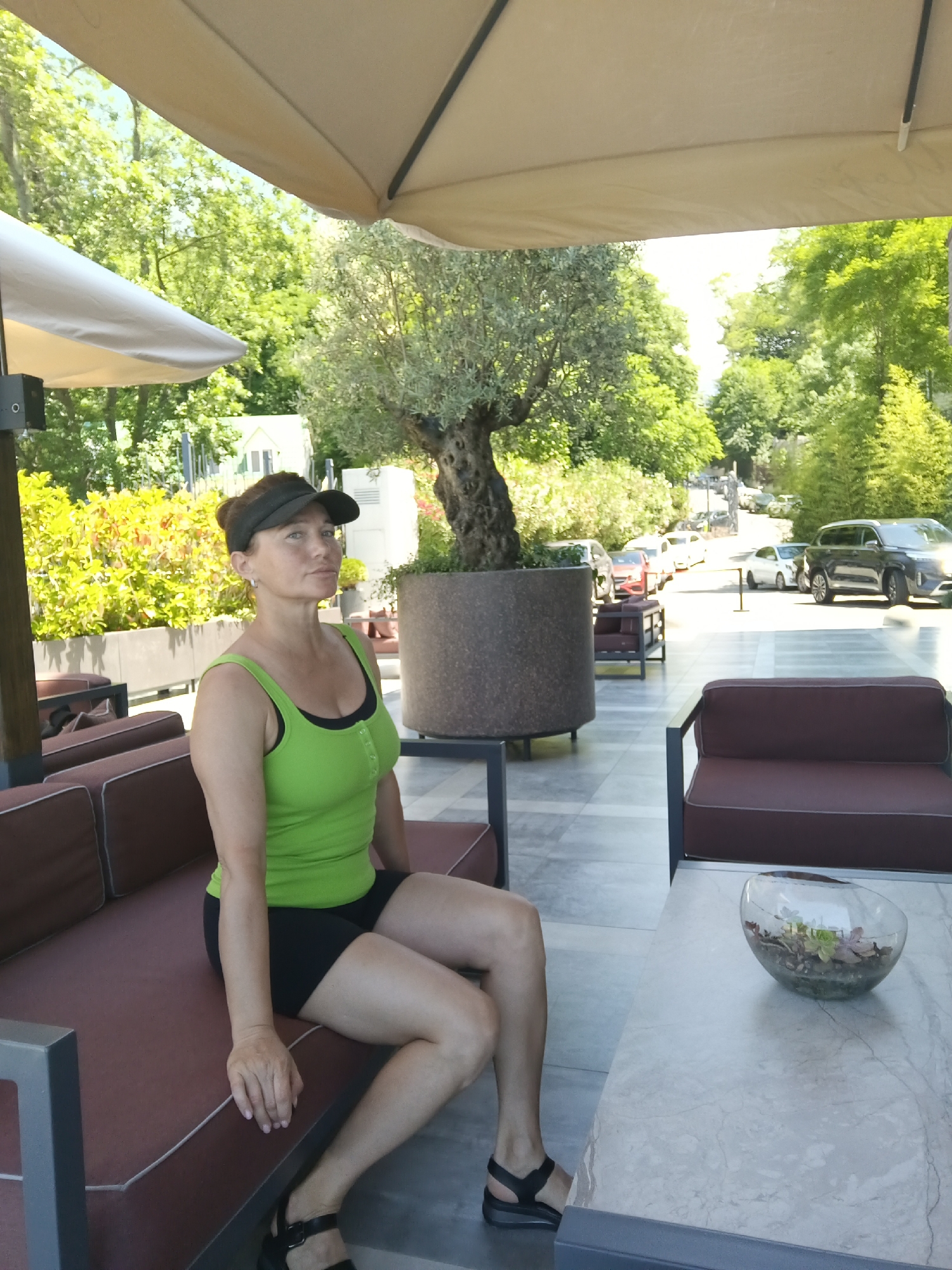 An outdoor seating area is depicted, with a woman sitting on a dark red cushioned sofa. She is wearing a lime green tank top and black shorts, and a black visor. She has her right arm resting on her lap and her left arm is partially visible. In front of the sofa is a light-colored marble table with a glass bowl containing small plants on it. Behind the woman, the background shows a large potted tree with an olive tree, a dense hedge of yellow and green foliage, and more trees and greenery. Several cars are visible on a road in the distance, and a large umbrella is shading part of the scene. The setting appears to be a restaurant or a cafe in a well-lit, outdoor environment with lush vegetation.