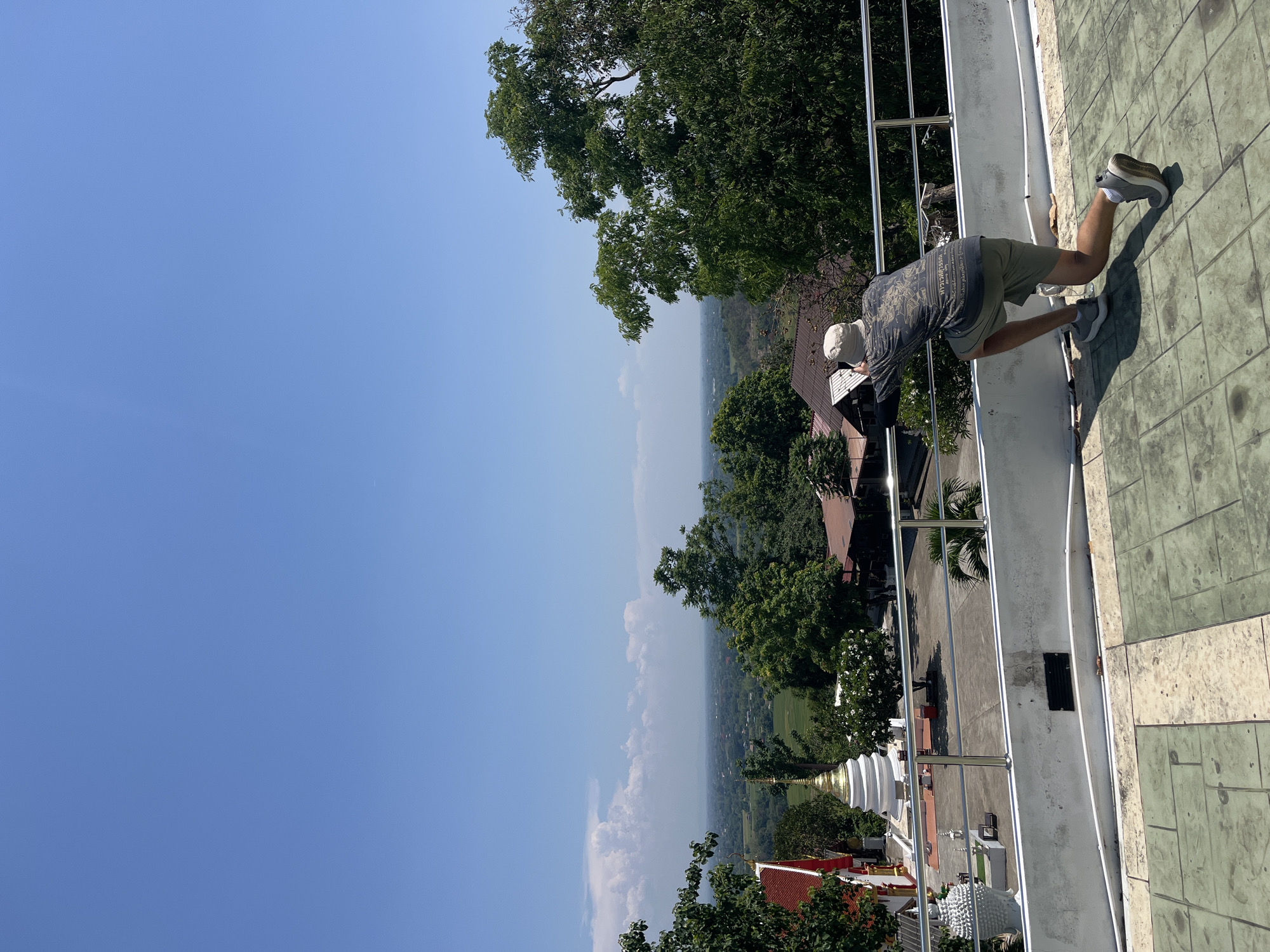 An outdoor photograph captures a man leaning over a railing, looking at a temple and scenic view in Nong Yaeng, Thailand. The man, positioned on the right side of the frame, wears a gray shirt with patterns, khaki shorts, gray sneakers, and a white hat. He is leaning forward, resting his hands on the silver metal railing.
In the background, a partially obscured view of a Buddhist temple is visible, featuring a white and gold spire. Beyond the temple, the vista opens up to a landscape filled with trees and glimpses of buildings, indicative of a cityscape or town nestled within a lush environment.
The sky occupies a significant portion of the image, a clear and bright blue, suggesting a sunny day. The color palette is predominantly composed of natural tones – blues, greens, whites, and browns – enhancing the impression of a scenic outdoor environment.