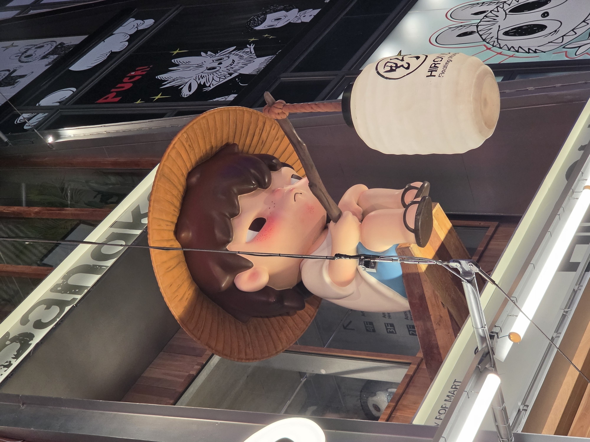 This image captures a large, suspended sculpture of a cartoon-style, child-like figure, likely part of an outdoor art installation or building facade in Bangkok, Thailand. The sculpture depicts a character with brown hair, fair skin, and prominent rosy cheeks, wearing a wide-brimmed, conical straw hat. It appears to be holding a dark, gnarled staff or stick across its body, from which a large, white, elongated lantern is suspended. The lantern features black text, including "HIRO Floating World" and some Japanese characters.
The figure is positioned against a background of a building facade, which includes multiple black panels adorned with white graphic illustrations of cartoon monsters and characters. One such panel clearly displays the word "FUCHI" in red lettering. Other parts of the building feature reflective surfaces and additional signs, with partially visible text like "V OF MART" (suggesting "POP MART") and floor indicators such as "3F" and "4F" accompanied by arrows. The sculpture is held in place by visible cables or wires.
The scene appears to be set at dusk or night, indicated by the illumination of the signs and the glow emanating from the lantern. The character's expression is gentle and serene, contributing to a whimsical and playful atmosphere within the urban commercial setting.