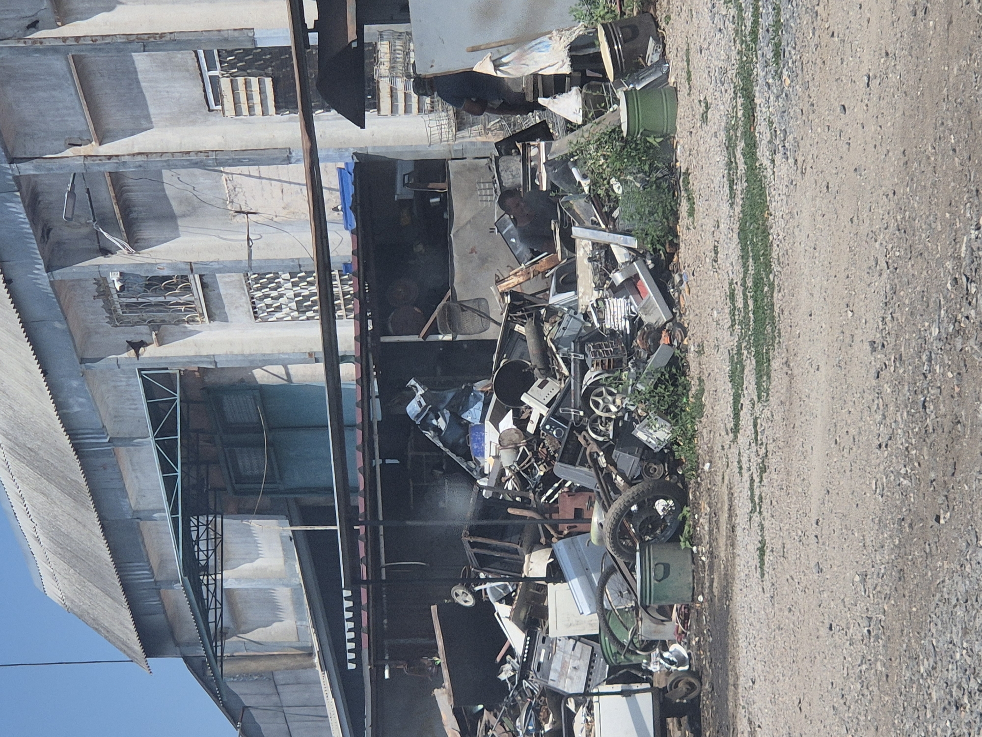 The image depicts an outdoor scene in what appears to be a storage or processing area for salvaged materials, possibly a scrap yard or recycling center. A large pile of various discarded objects, including electronics, metal parts, and other debris, is a prominent feature. Among the clutter, one can identify components of computers, printers, wheels from various vehicles, and metal casings. Some of these items are stacked precariously, while others are scattered across the ground.
Behind the pile of scrap, a multi-story building with a weathered, possibly concrete facade is visible. It has several windows, some of which are barred or covered with patterned screens. The building's architecture suggests it could be an industrial or commercial structure, or perhaps an older residential building. Adjacent to the scrap pile, there are also some green barrels and plants, indicating an outdoor environment with vegetation. A rough, gravelly path or road runs along the right side of the image, bordering the area where the scrap is accumulated.
The lighting suggests it is daytime, with sunlight casting shadows. The weather appears to be fair. There are no distinct signs of specific activity or event beyond the general state of disarray and accumulation of discarded items. No visible text is discernible in the image. The overall impression is one of a functional but somewhat unkempt outdoor industrial or salvage area.