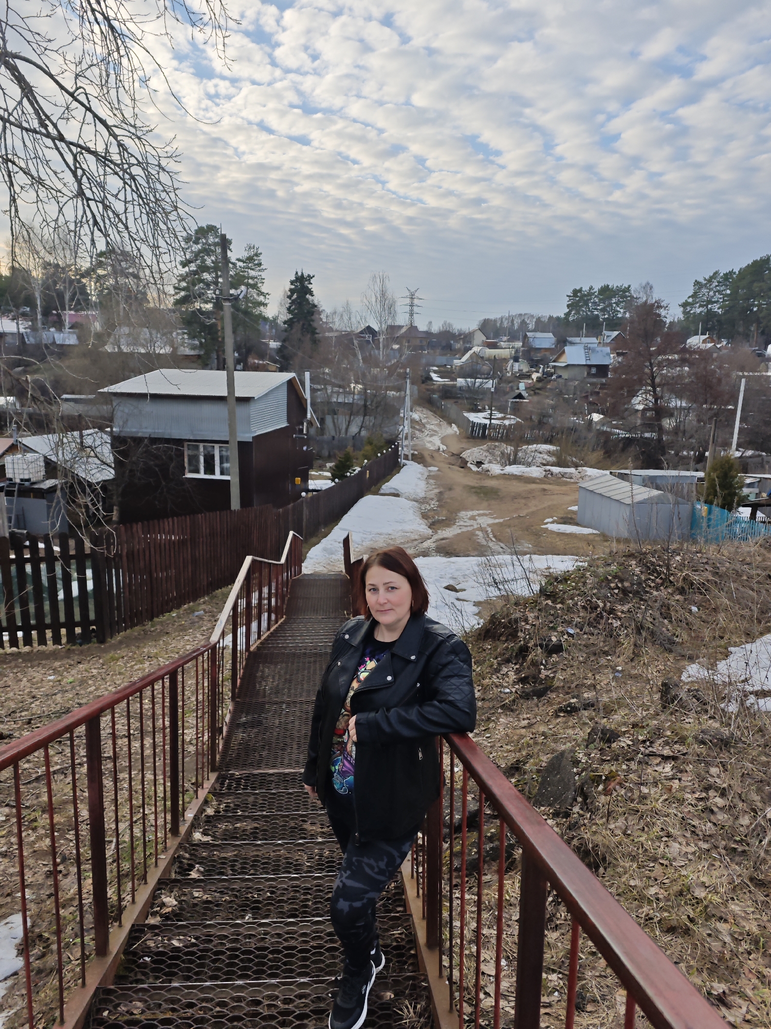 A woman stands on metal stairs with red railings in Chaikovsky, Russia. The stairs lead down from a residential area with small houses and trees, with patches of snow visible on the ground. The sky is overcast with patterned clouds. The woman is wearing a black leather jacket, a colorful shirt, patterned leggings, and black sneakers. She is leaning against the railing and looking at the camera. The scene appears to be early spring.