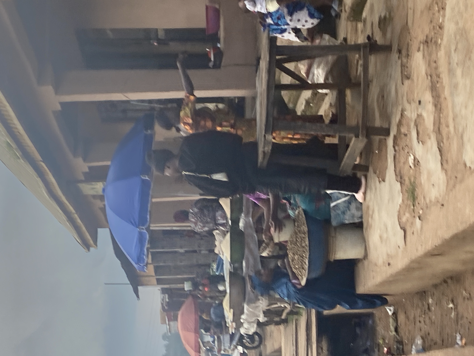 The image depicts an outdoor market scene in Bukuru, Nigeria, under an overcast sky. In the foreground, a woman in a blue garment is partially visible, attending to a large basin filled with small, brown items, possibly grains or nuts. Beside her, a white bucket sits on the ground. A wooden table with a rustic design is positioned in the mid-ground, its surface cluttered with various items. Behind the table, two individuals are engaged in activity; one, wearing a black outfit and headscarf, stands facing left, while another in a vibrantly patterned dress gestures with their right arm. A large blue umbrella provides shade over a stall. In the background, more stalls with umbrellas and wares are visible, along with a red umbrella, a motorcycle, and other market-goers. The ground is a mix of dirt and concrete, with scattered debris. The architecture of the buildings in the background appears simple, with covered walkways.