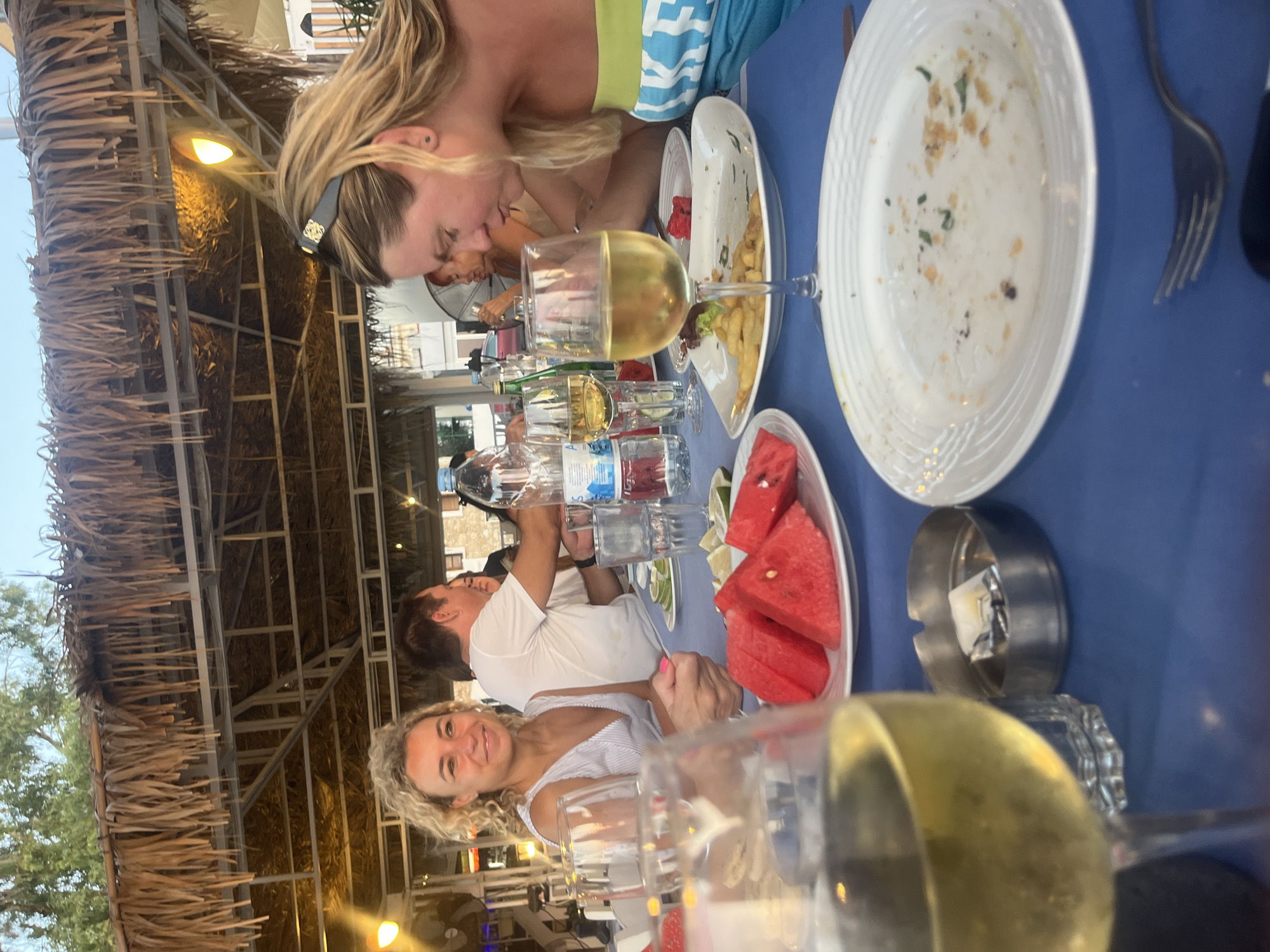 The image captures an outdoor dining scene at a restaurant in Polis Chrysochous, Cyprus, possibly a taverna given the thatched roof. The setting appears to be late afternoon or early evening, suggested by the warm lighting and the sky visible through the roof structure. A blue tablecloth covers the table, which is laden with remnants of a meal.
In the foreground, a glass of white wine is prominently featured, slightly out of focus. To its right, an ashtray with a cigarette butt and a lighter is visible. Further back, a white plate with leftover food, including french fries and some green garnish, sits before a glass of white wine. To the left, a plate of sliced watermelon and lime wedges indicates fresh fruit served.
Three individuals are partially visible at the table. A woman with blonde, curly hair and a white, striped top is seated on the left, smiling. Next to her, a man in a white t-shirt is also seated, looking to his right. On the right side of the frame, another woman with long blonde hair and a blue and green strapless top is leaning forward, looking down at her plate. The overall atmosphere suggests a relaxed meal shared by friends or family. The background shows more diners and the open-air structure of the restaurant.