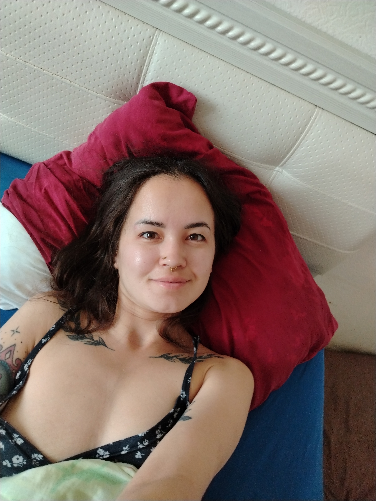 A woman is lying on a bed. She is wearing a black floral patterned top. She has tattoos on her chest and upper arm. Her head is resting on a red pillow. There is a white pillow to her left. The bed has a blue sheet. The headboard of the bed is white with a decorative trim. The location is unknown, and cannot be discerned as Noviy, Russia, based on the image.