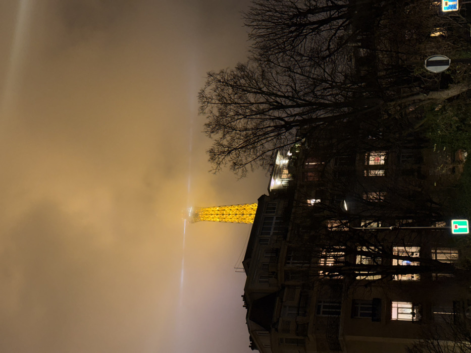 The Eiffel Tower glows yellow through a thick fog, appearing as a narrow beacon. A residential building with many lighted windows sits on the right side of the image. Bare branches from several trees partially obscure the buildings and the sky is filled with an orange hue due to the fog. A street lamp and road signs are also visible along the roadside. The image is oriented sideways.