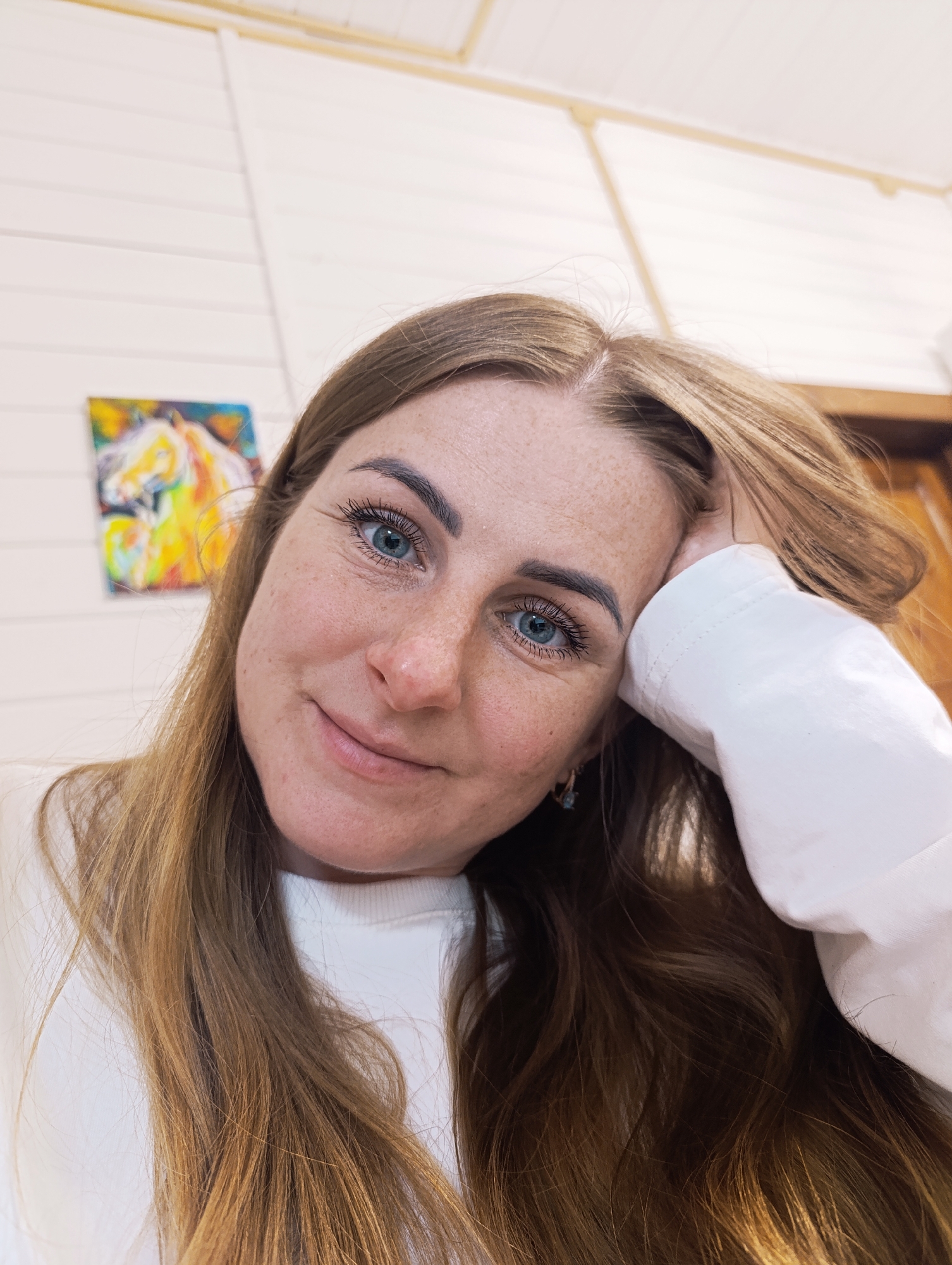 A close-up photograph presents a woman indoors, possibly in a residential setting in the city of Unknown, Unknown. She is fair-skinned with freckles and light brown hair, wearing a white long-sleeved shirt. Her left hand is resting on her head, and she has a slight smile. A painting of a horse in bright colors is visible on the wall behind her. The wall is paneled and painted white. A dark wooden door or frame is partially visible on the right side of the image.