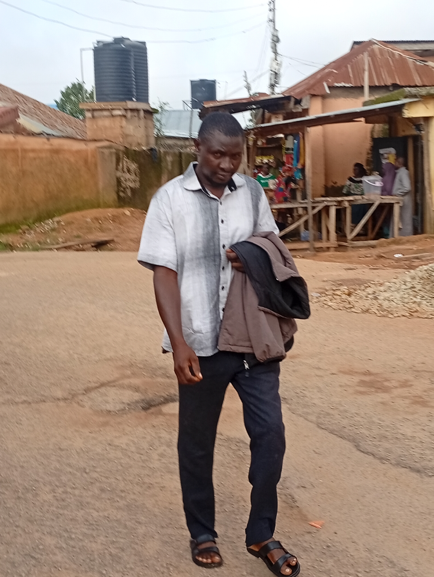 The image depicts a street scene in Jos, Nigeria, likely in a commercial or residential area characterized by modest buildings and an unpaved road. In the foreground, a dark-skinned man wearing a gray and white striped short-sleeved shirt, dark trousers, and black sandals is walking towards the camera. He is carrying a folded brown and black jacket over his left arm. The man appears to be looking directly at the camera.
In the background, several structures are visible. To the left, a large black water tank sits atop a building. Further to the right, there is a small market or shop area with a corrugated iron roof and wooden stalls. Some people are standing or sitting within this area, suggesting ongoing commercial activity. The overall environment appears somewhat dusty, with scattered debris on the ground, indicative of a typical street in a developing urban setting. The sky is overcast, contributing to a diffused light quality in the photograph.