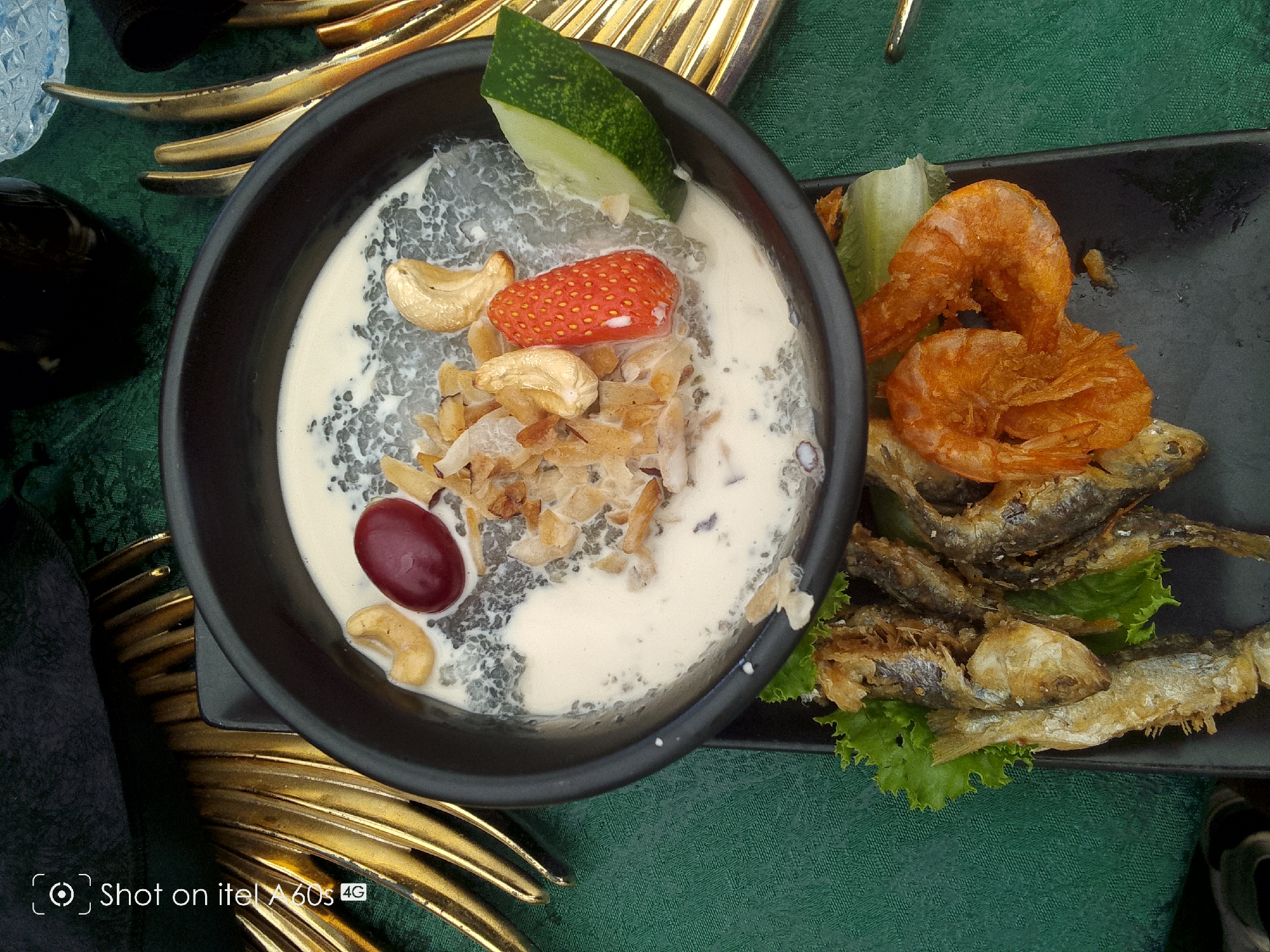 The image depicts a meal with a bowl of dessert and a plate of seafood. The dessert is in a black bowl containing a creamy base with chia seeds, topped with slices of strawberry, grape, cucumber, cashews, and toasted coconut flakes. Adjacent to the bowl is a black plate with fried shrimp and small fried fish on a bed of lettuce. The setting includes gold decorative objects and a green textured surface resembling a tablecloth. The image was captured using an itel A60s mobile phone. The location is specified as Idimu, Nigeria.