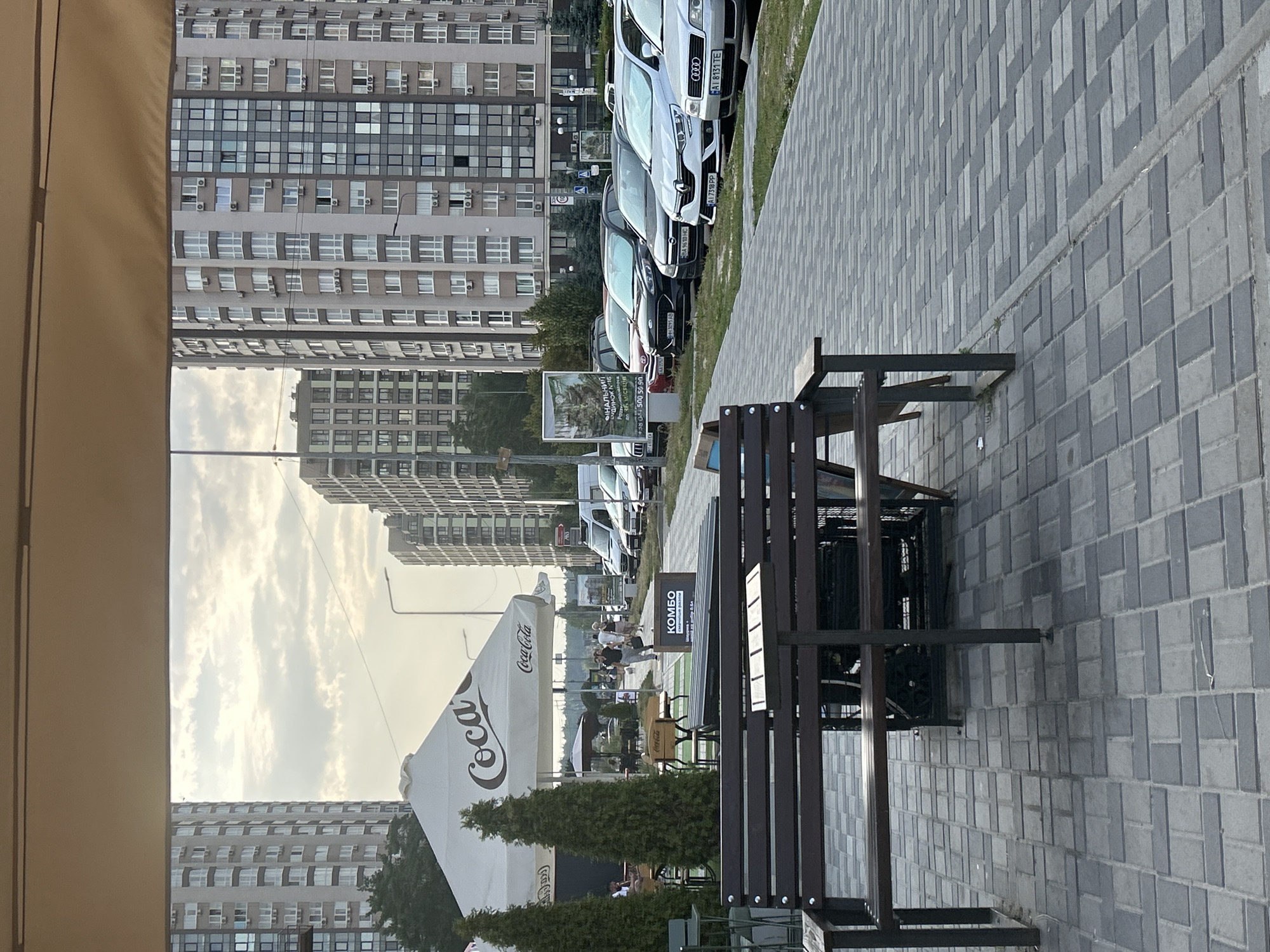 The image depicts an outdoor seating area with parked cars and apartment buildings in the background, suggesting an urban setting. In the foreground, there are dark brown wooden benches and tables on a grey paved walkway. To the left, there is a large white Coca-Cola umbrella and two tall, conical evergreen trees. Further back, people are seen walking and sitting at tables under umbrellas. A row of parked cars lines the right side of the paved area, with several multi-story apartment buildings visible behind them. A sign with Ukrainian text, including "КОМБО," is visible. The sky is overcast with some clouds. The general atmosphere appears to be that of a commercial or residential area in Brovary, Ukraine, likely a cafe or restaurant patio adjacent to a street with parking and residential buildings.