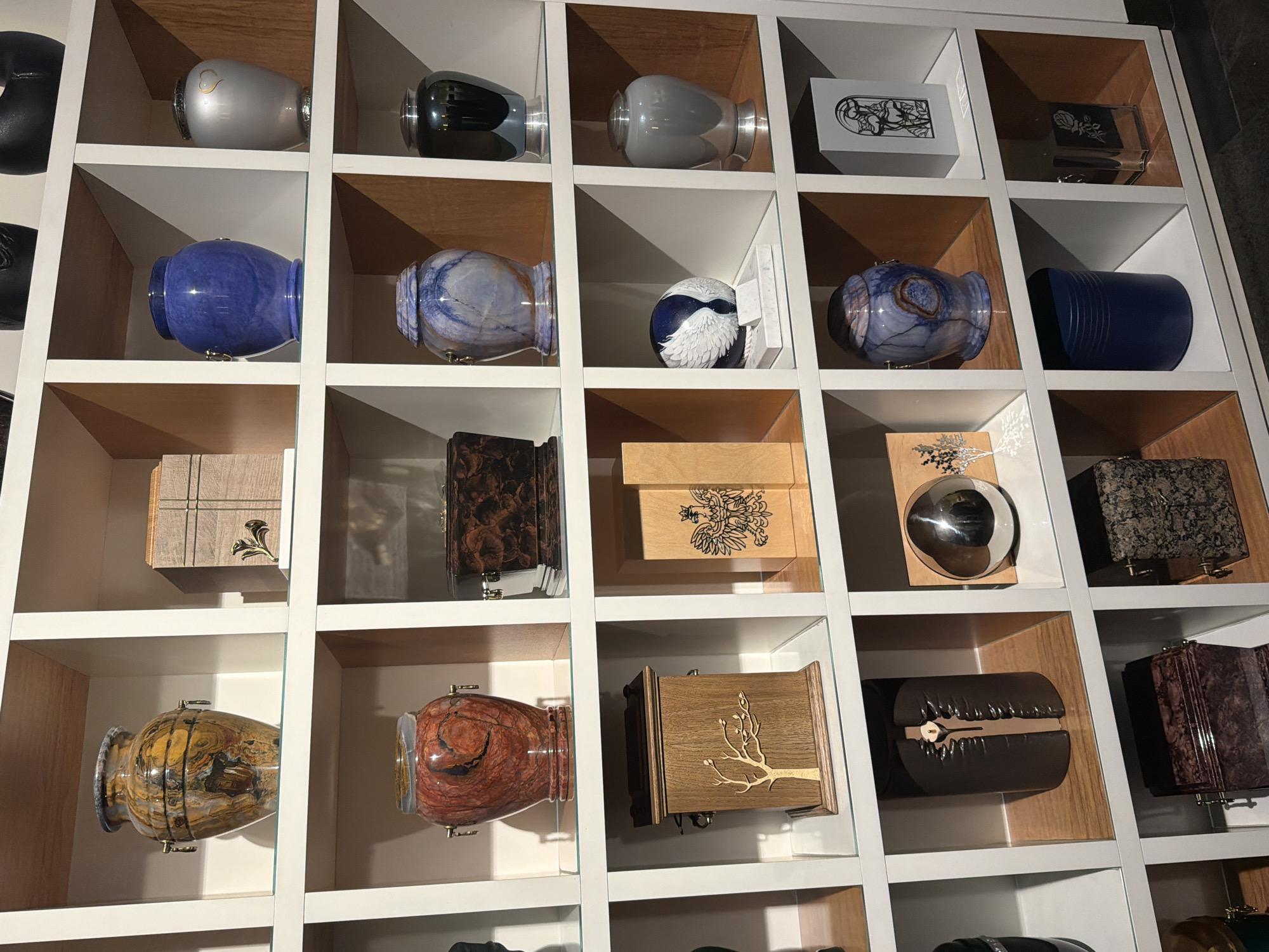 The image depicts a display of urns, likely for ashes, within a white shelving unit with wooden backings in a location in Unknown, Unknown. The urns are of various styles and materials, including ceramic, wood, and metal, and are arranged in rows within the individual cubbies of the shelving unit. There are no people visible in the frame. The focus is on the assortment and presentation of the urns.