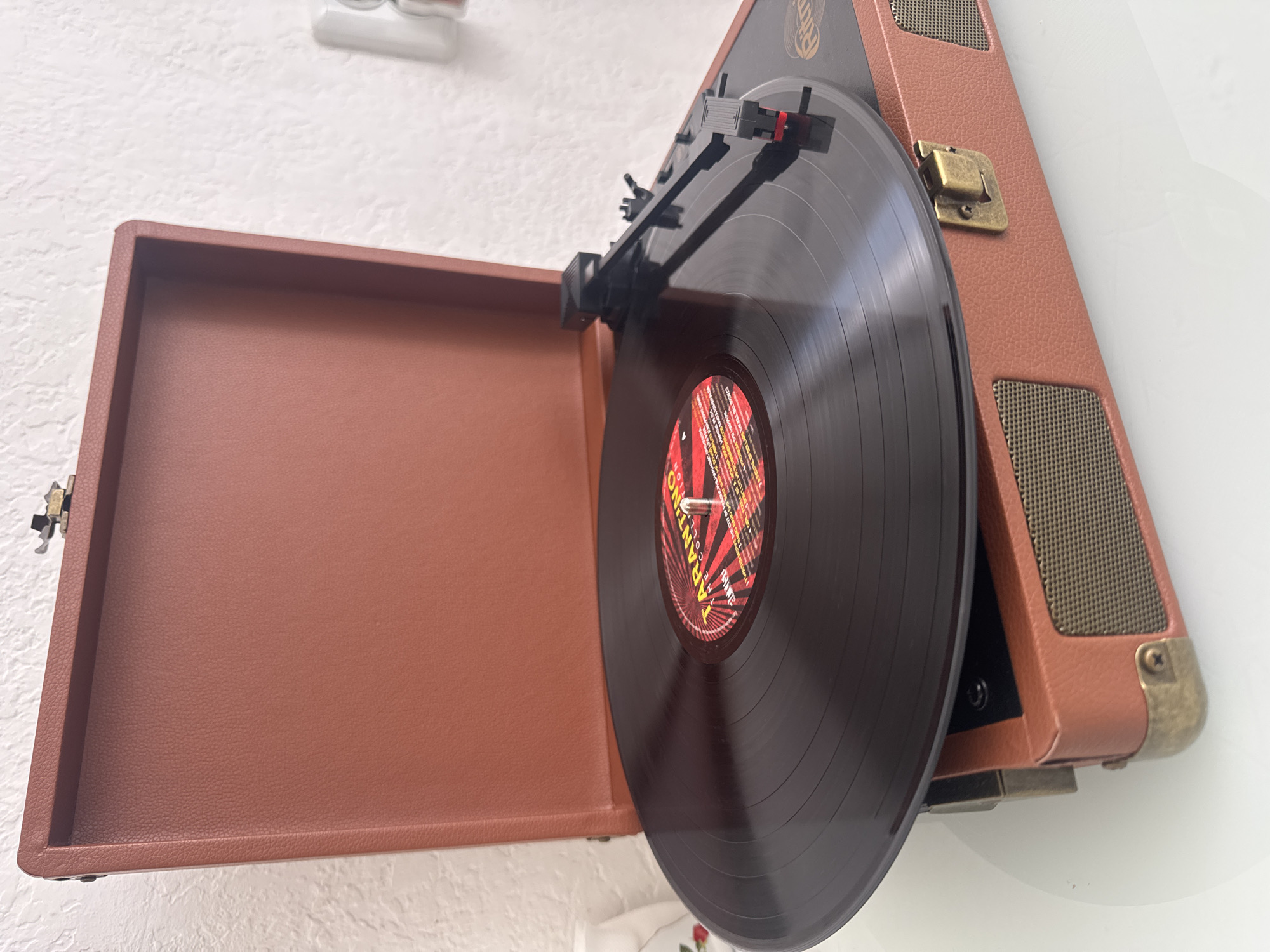 The image shows a close-up of a vintage-style record player with a vinyl record on it. The record player is open, revealing the turntable and tonearm with a needle. The record itself has a colorful label. The record player is made of a brown material, possibly faux leather, with metal accents. The background is a textured white wall. The scene appears to be indoors, on a white surface. There is no indication of the city of Ufa, Russia, or any specific location. The image is a still photograph.