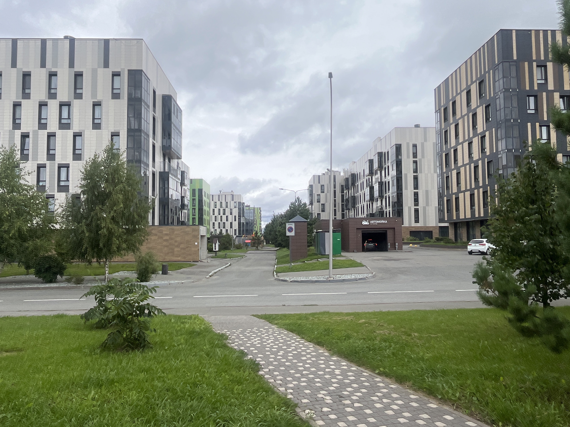 The image shows a street view in Innopolis, Russia, with modern multi-story buildings lining both sides of the road. The buildings feature a contemporary architectural style with varied facade designs, including vertical stripes of different colors and materials. On the left, a light-colored building with gray and black accents is prominent, with several green buildings visible further down the street. On the right, a building with dark gray and tan vertical stripes is in the foreground, with another light-colored building behind it.
In the middle ground, a road curves through the scene, with white lane markings. Several trees and green spaces are present, including a paved walkway with a patterned surface in the lower left of the frame. To the right of the road, there is a car wash facility with "Автомойка" (Car Wash) signage. A white car is parked in front of the car wash, and a black car is visible inside the bay. A street light stands tall in the distance. The sky is overcast with clouds. There are no people clearly visible in the image.