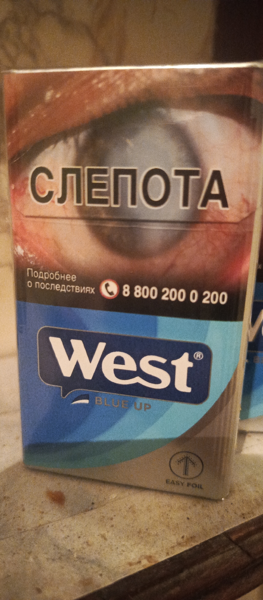 This is a close-up, slightly angled photograph of the front of a cigarette pack. The pack features a stark image of an eye with what appears to be a cataract, conveying a warning. In large, bold, black letters, the Russian word "СЛЕПОТА" (meaning "blindness") is prominently displayed across the top portion of the pack, overlaying the eye.
Below this warning, smaller text in Russian reads "Подробнее о последствиях" (meaning "More about the consequences"). To the right of this text, a phone icon is visible next to the number "8 800 200 0 200".
The lower half of the pack is dominated by a blue color scheme. A prominent white logo, "West", is presented in a speech bubble-like graphic. Beneath the "West" logo, the words "BLUE UP" are printed in a lighter blue. In the bottom right corner of the pack, a small graphic of an arrow pointing upwards within a circle is visible, with the text "EASY FOIL" printed below it.
The lighting in the image appears to be artificial, casting a warm tone on the surrounding surface, which seems to be a textured, possibly stone or wood, countertop. The background is out of focus but suggests an indoor setting. The overall mood conveyed by the pack design is serious and cautionary, due to the prominent warning about blindness. The context of Polyarnye Zori, Russia, indicates the geographical origin of the product.