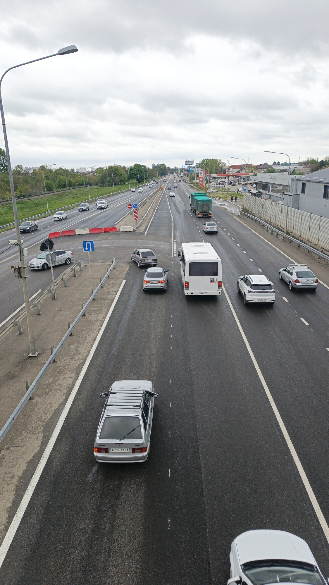 The image depicts a multi-lane highway in Krasnodar, Russia, under cloudy skies. The road surface appears wet. Several vehicles are visible including cars, a bus, and a truck. The vehicles are traveling in both directions on the highway. Roadside features include guardrails, lane markings, light poles, vegetation, buildings, and road signs. Red and white traffic barriers are present near a U-turn sign.