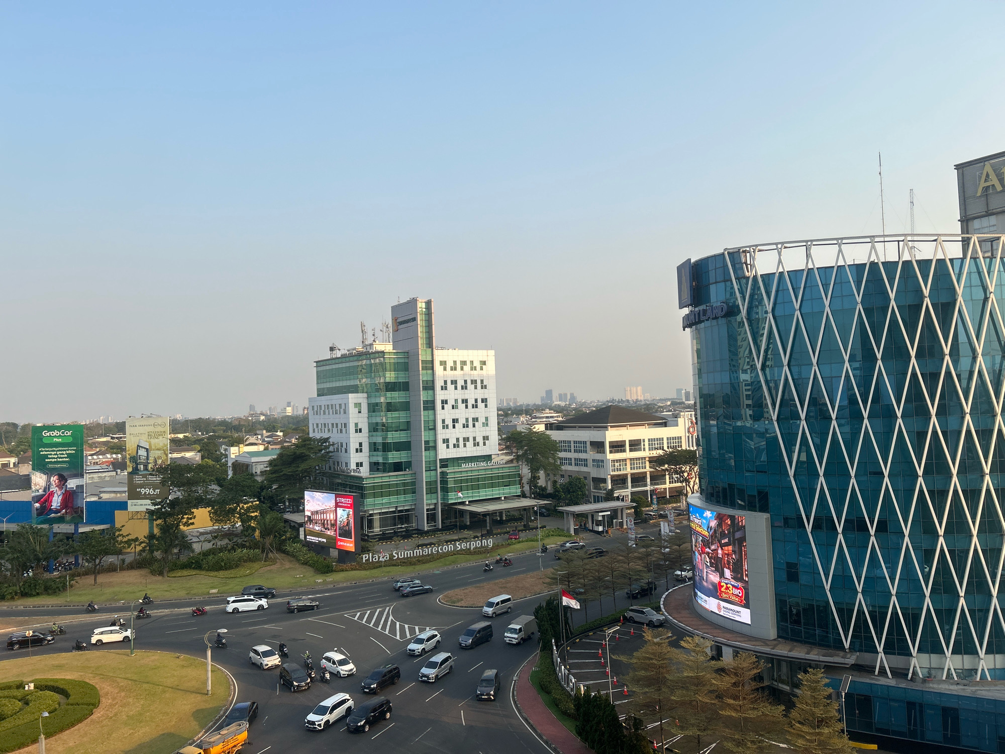 The image captures an aerial view of a busy intersection in Pakulonan Barat, Indonesia, likely within a commercial district, bathed in the soft light of late afternoon. A prominent feature is the Plaza Summarecon Serpong, a modern building with a green facade and white geometric patterns. Adjacent to it is another contemporary glass-encased building with a diamond-shaped pattern on its exterior, and a large digital billboard displaying advertisements for "Strozzi" and "Paramount Land." The intersection itself is a hive of activity, with numerous vehicles including cars, vans, and motorcycles navigating the road. Several individuals are visible on motorcycles. The surrounding landscape includes other commercial buildings, trees, and a well-maintained grassy roundabout. In the background, a hazy cityscape extends towards the horizon. A billboard advertising "GrabCar Plus" is visible on the left. The sky is a clear, pale blue, indicating a generally fair weather condition.