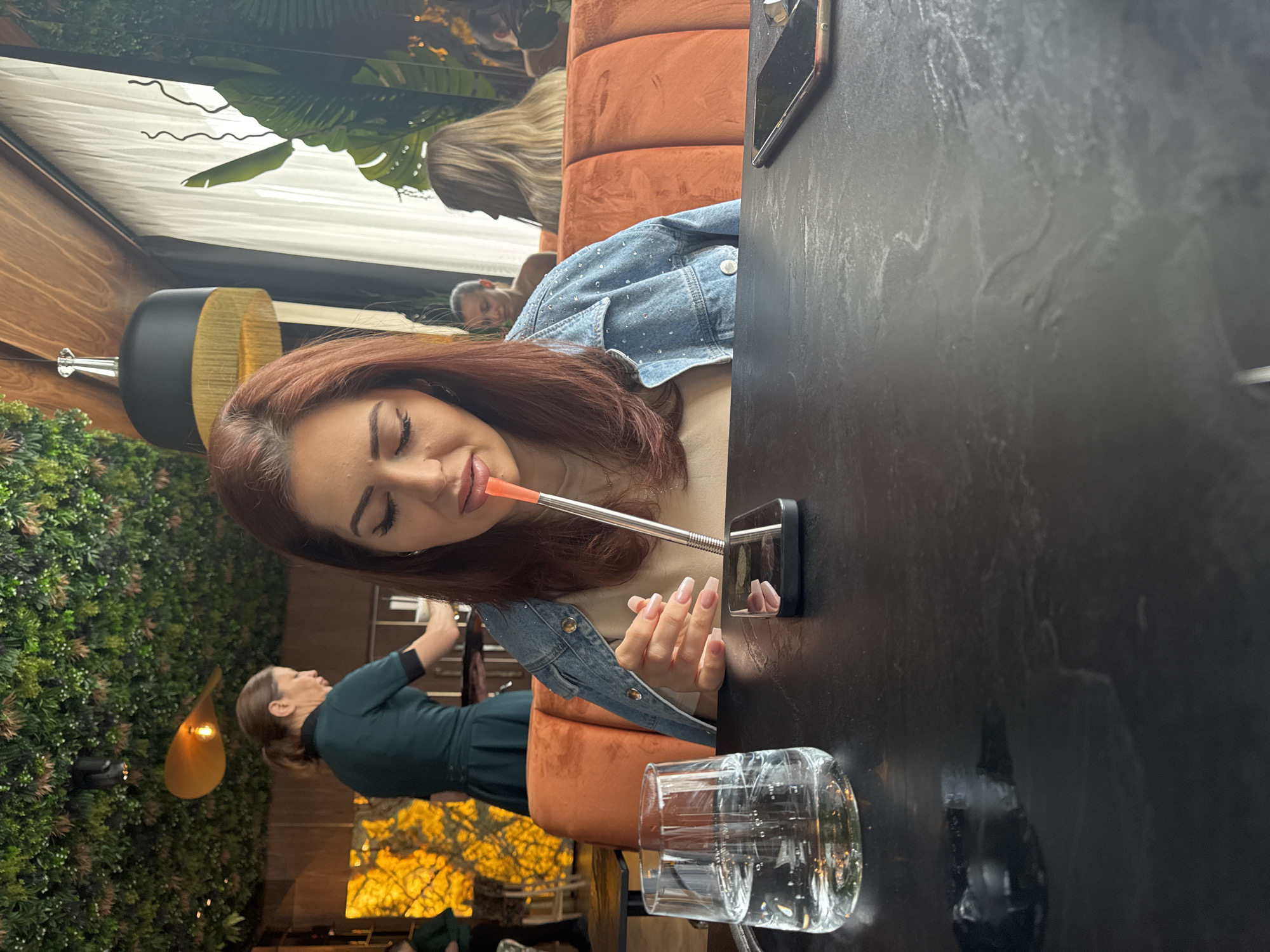 A woman with auburn hair is sitting at a table indoors, possibly in Sofia, Bulgaria. She is wearing a denim jacket and is using a metal straw to drink from a glass. Other people are visible in the background, including a woman in a green dress and another woman with blonde hair. The decor includes greenery and lamps.