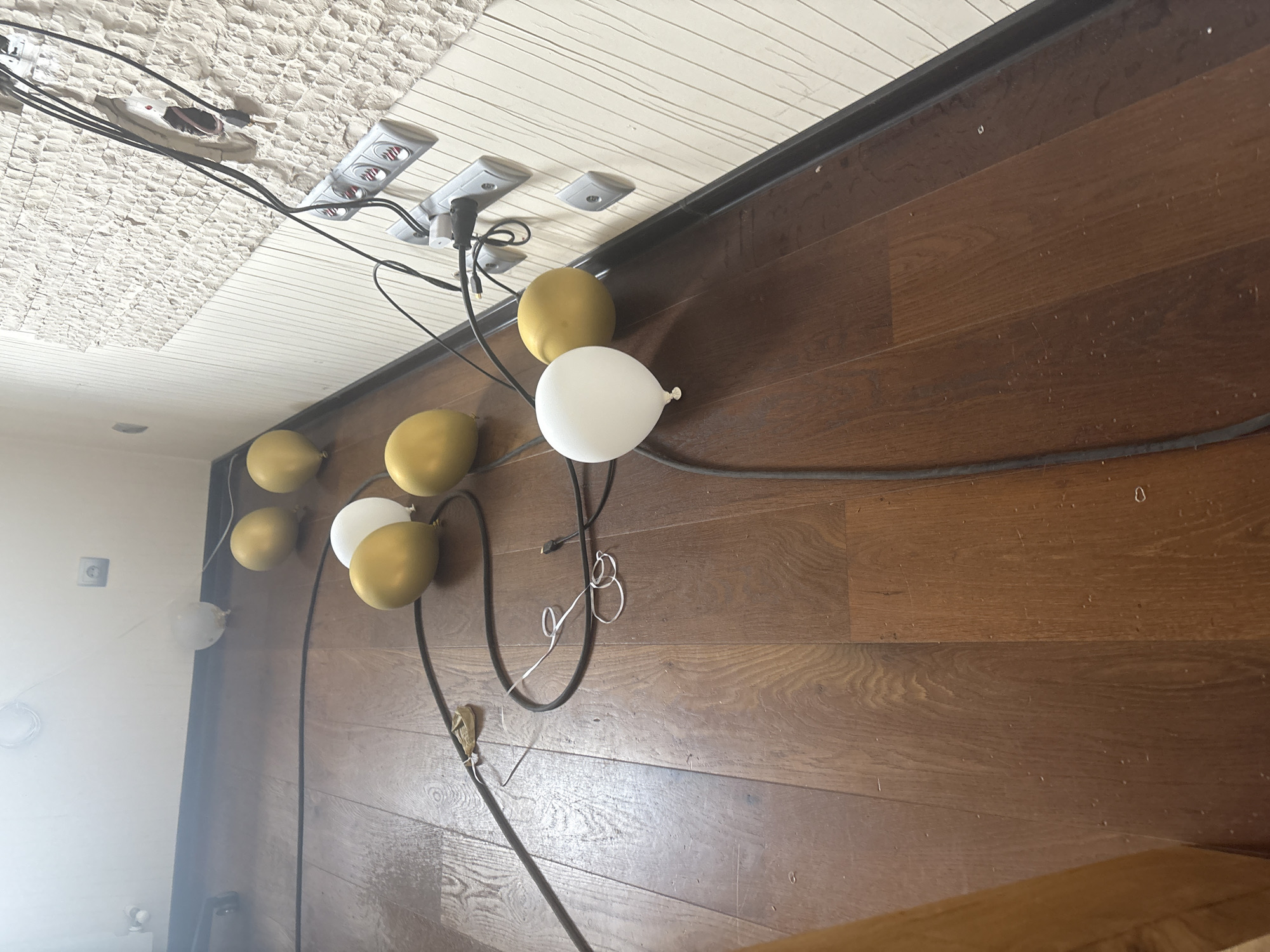 The image shows a close-up of a wooden floor with several balloons scattered around. Some of the balloons are gold, while others are white. There are also electrical cords and power outlets visible on the wall in the background. The scene appears to be indoors, in a room with white walls and a textured surface. The overall impression is one of a partially decorated or deconstructed event space, possibly a party or celebration that has recently concluded or is in preparation. The lighting is bright, with some areas appearing slightly overexposed. There are no people visible in the image. The location is specified as Orekhovka, Russia.