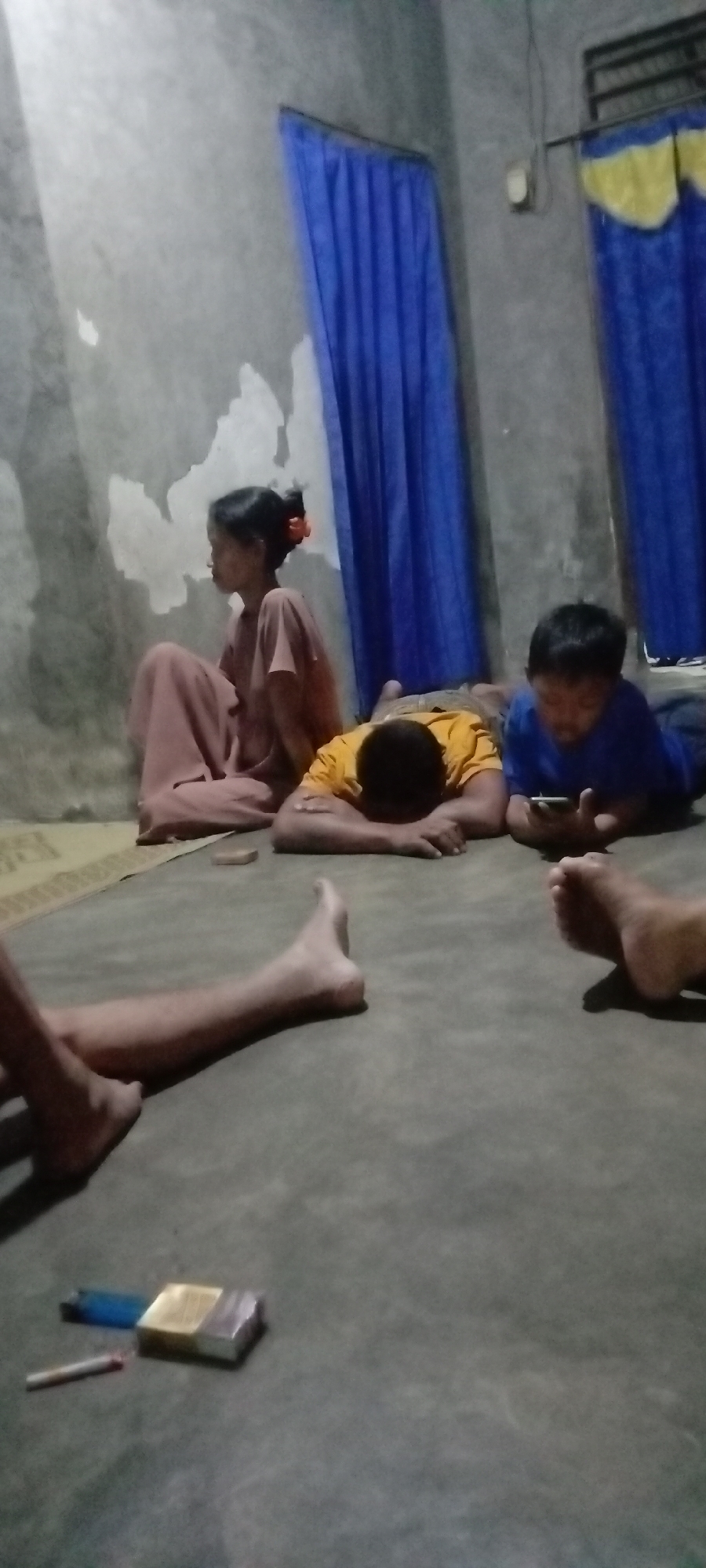 The image depicts a dimly lit room with a concrete floor and textured walls, likely a residential space in Utama Jaya, Mataram, Indonesia. Four individuals are present. A woman in a pink garment sits against the wall, her head turned to her left. Two children are lying on the floor. One child, wearing a yellow shirt, has their head resting on their arms, facing downwards. Another child, in a blue shirt, is looking at a mobile phone. The legs of a fifth person are visible in the foreground, and to their left, on the floor, are a cigarette pack, a lighter, and a cigarette. Blue curtains hang on the walls.