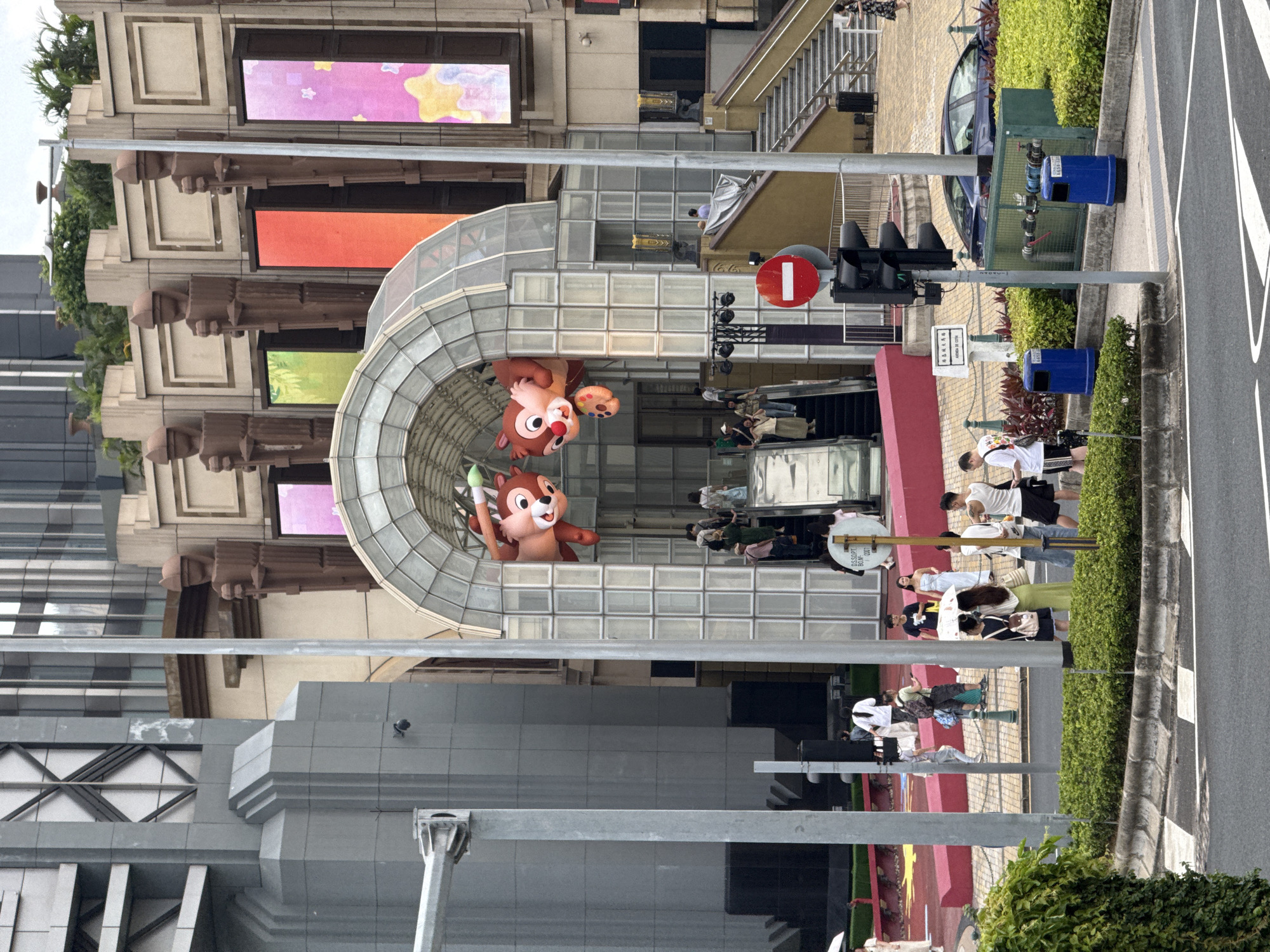 The scene shows a building exterior with a large Chip and Dale character figure. Pedestrians are walking by the building. The location is identified as Unknown, Macao. The weather appears to be clear. There is a traffic light at the side of the building.